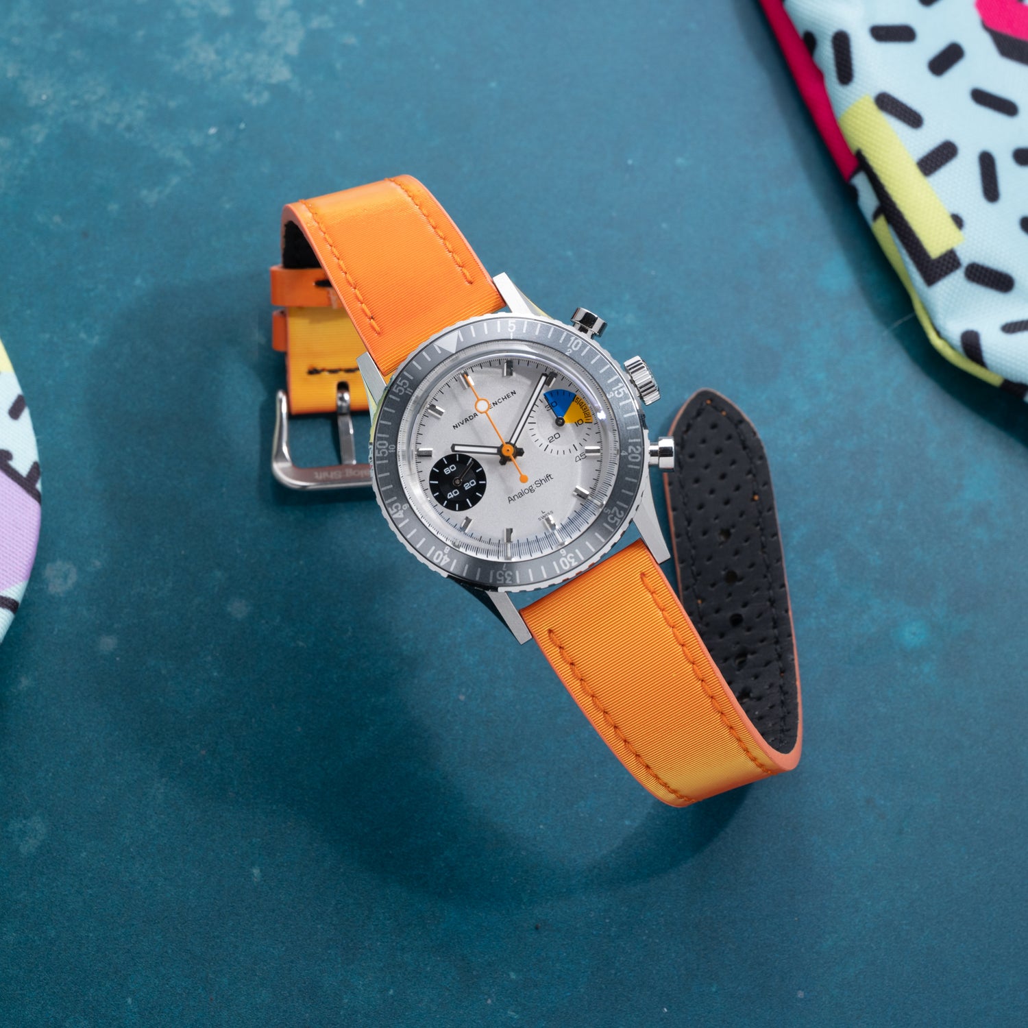 An Analog:Shift watch with a white dial, orange second hand, and limited Grabber Orange Effect Matrix Strap rests on a textured blue surface, surrounded by colorful patterned items.