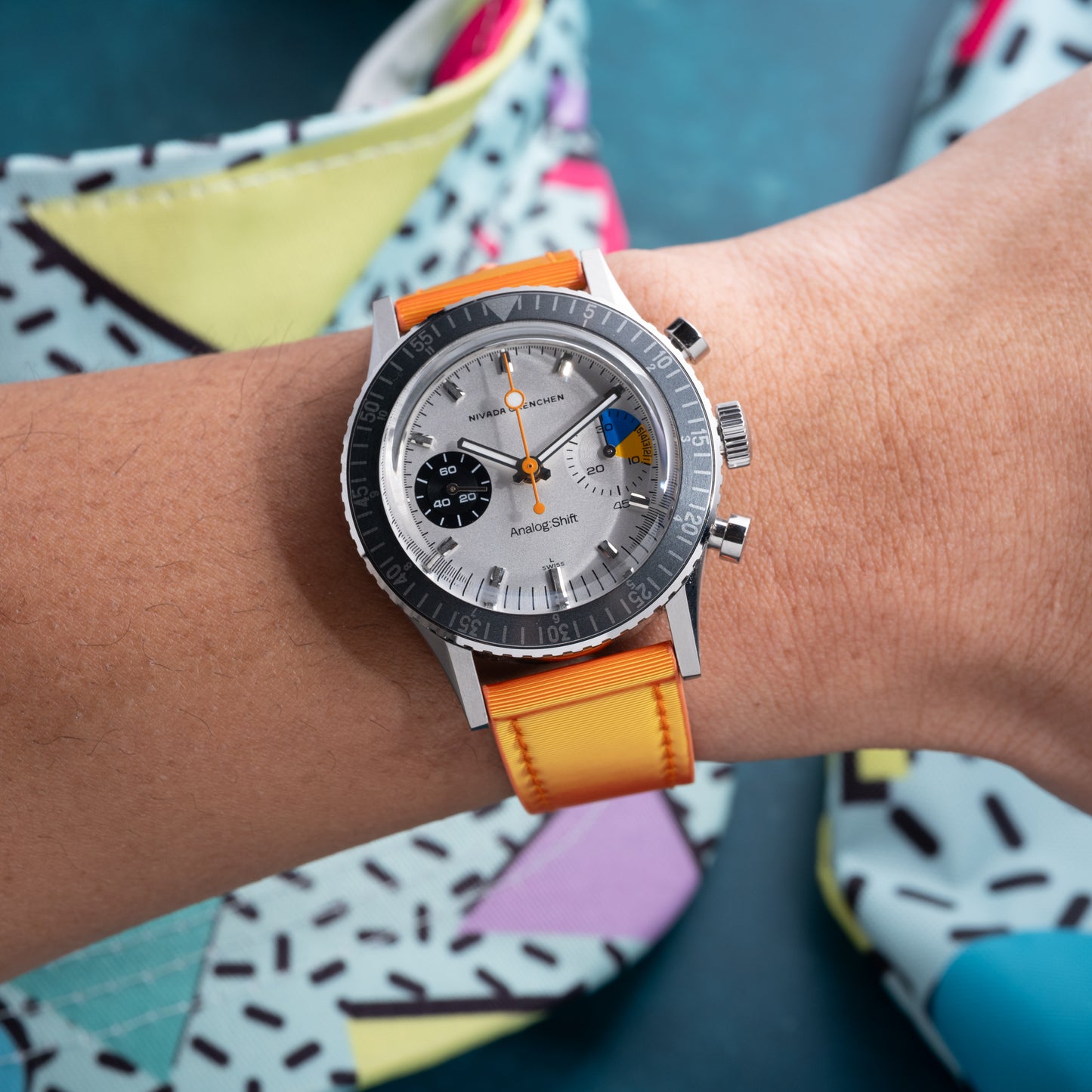 Someone wears a silver chronograph wristwatch with an Analog:Shift Grabber Orange Effect Matrix Strap. The watch has a white dial with black and orange accents, set among colorful geometric-patterned objects.