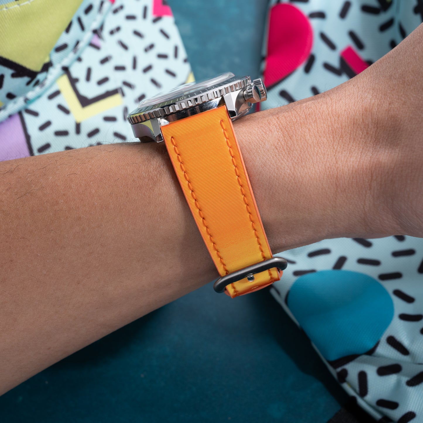 A close-up of a wrist wearing the Analog:Shift Grabber Orange Effect Matrix Strap, set against a colorful background with geometric and abstract shapes.