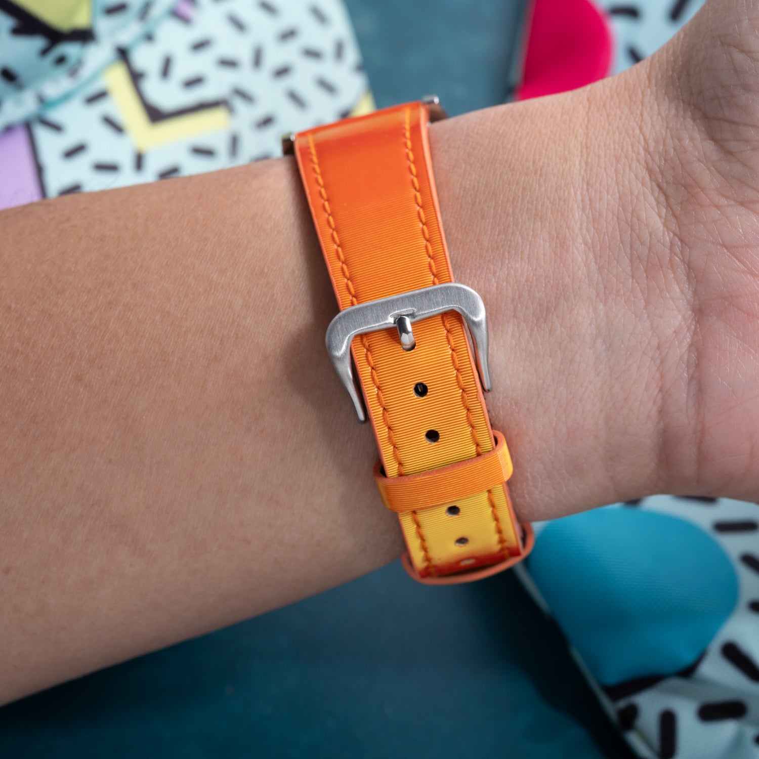 A close-up of a wrist wearing the Analog:Shift Grabber Orange Effect Matrix Strap with a silver buckle, set against a background of colorful abstract geometric shapes and bold lines.