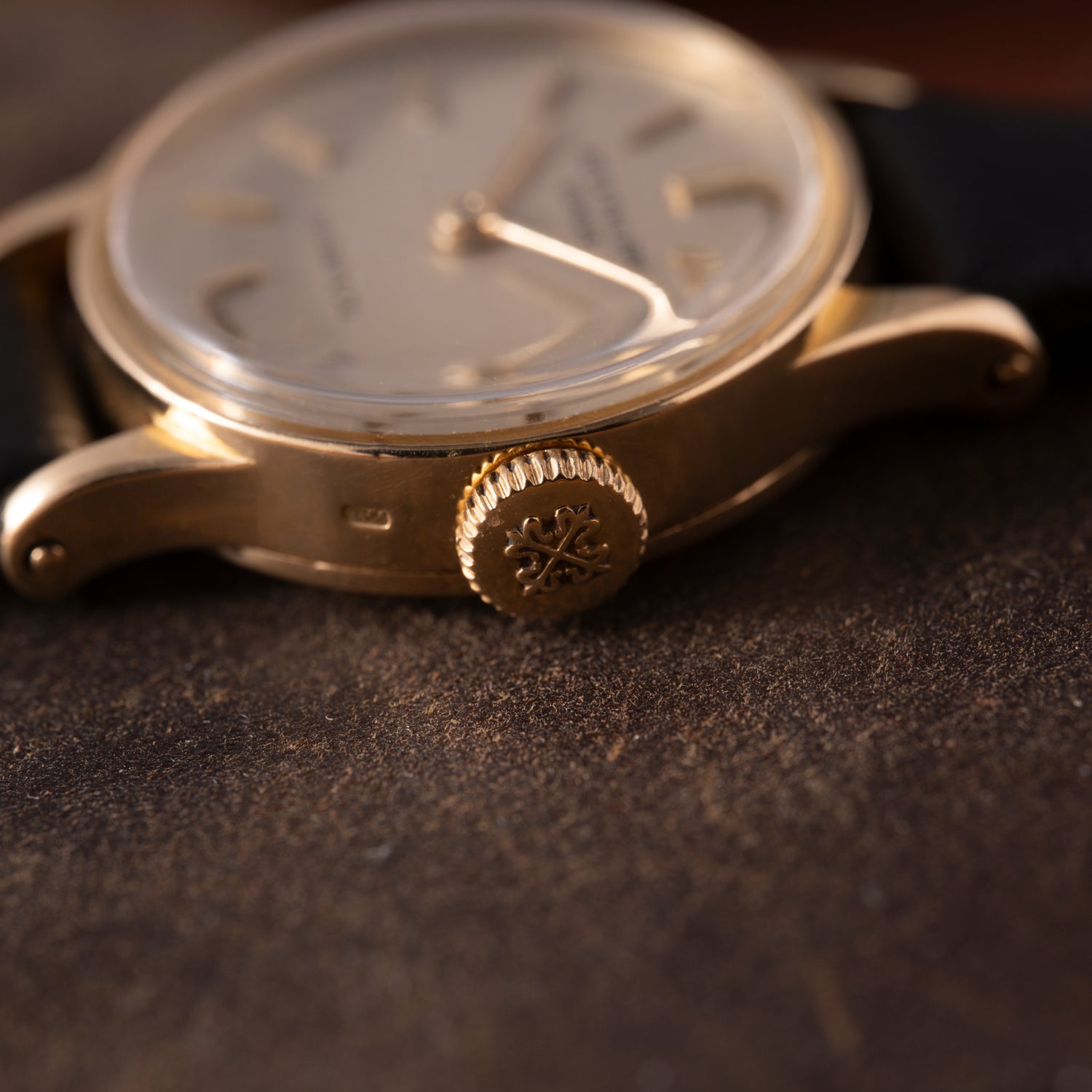 Close-up of the crown and side case of a Patek Philippe Calatrava 'Tiffany & Co' ladies dress watch in 18K yellow gold with a black strap, resting on a brown textured surface. The crown features an engraved floral design.