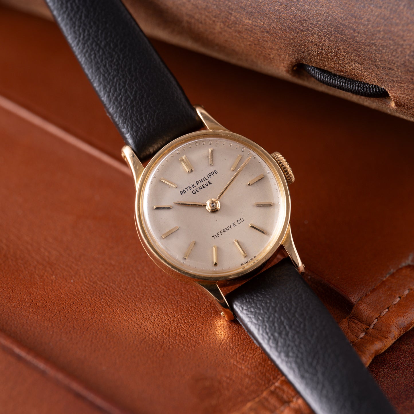 A Patek Philippe Calatrava 'Tiffany & Co' ladies dress watch with an 18K yellow gold case, white dial, and black leather strap sits on a brown leather surface near a notebook. "Patek Philippe Genève" and "Tiffany & Co." appear in gold on the dial.