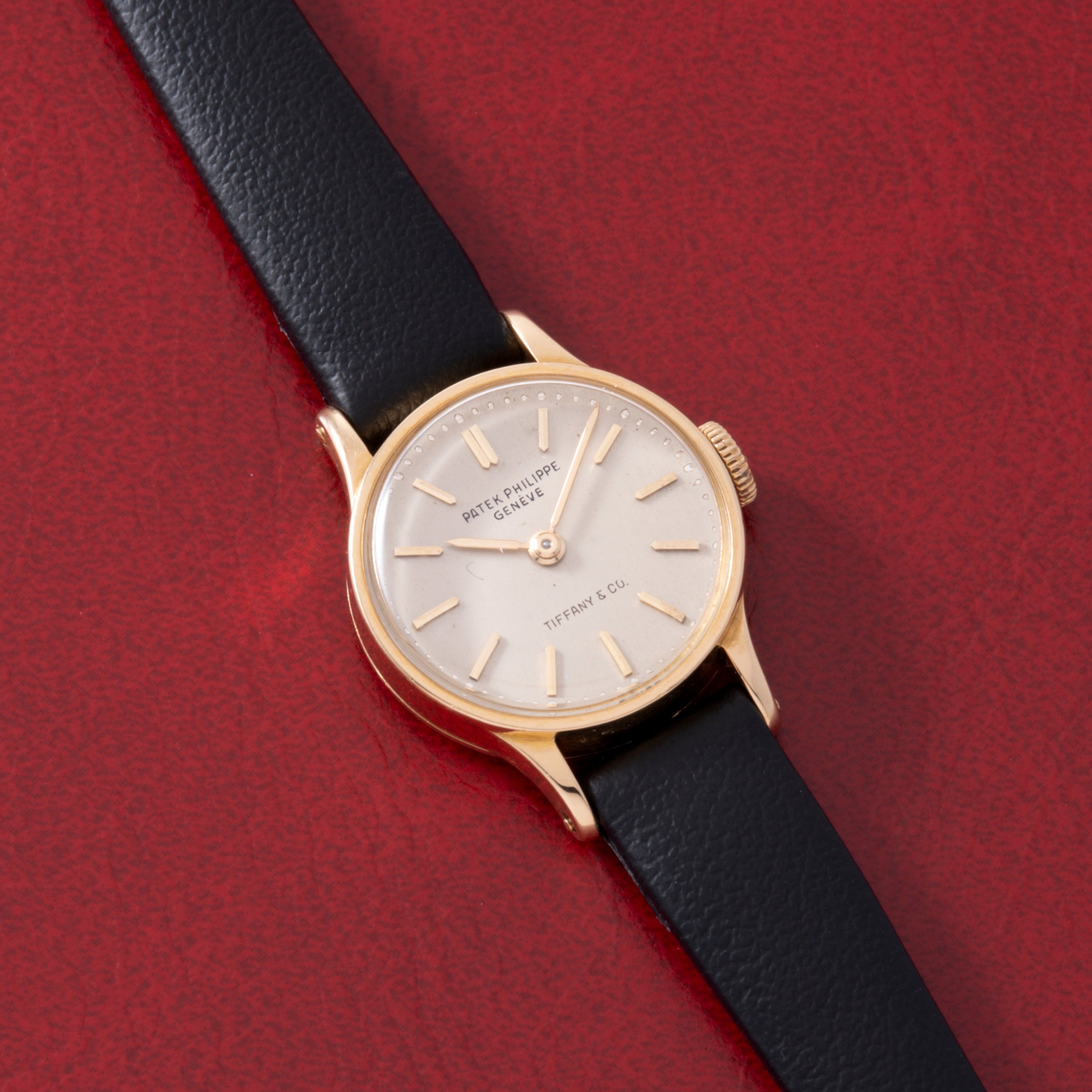 A Patek Philippe Calatrava 'Tiffany & Co' ladies dress watch in 18K yellow gold with a white dial, black leather strap, and gold hour markers is shown on a textured red surface. The dial displays Patek Philippe and Tiffany & Co.