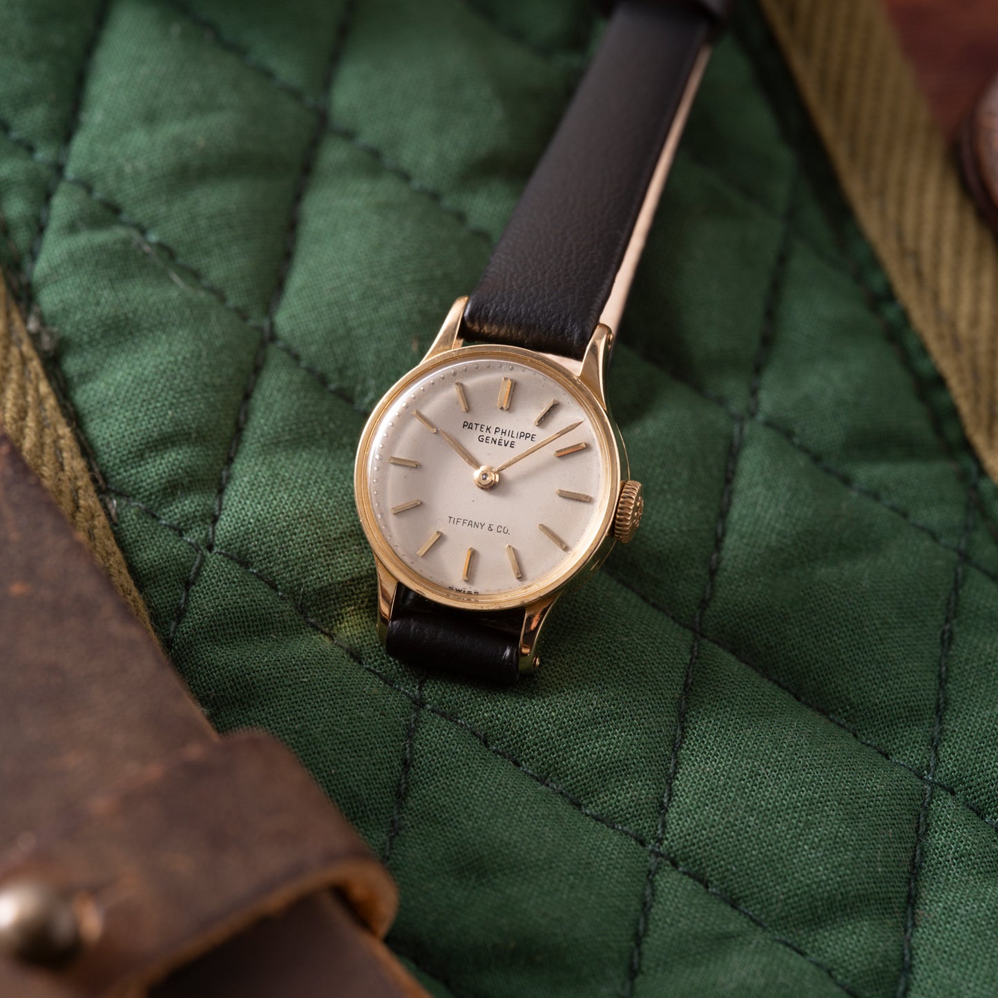 A Patek Philippe Calatrava 'Tiffany & Co' ladies watch with a black leather strap and 18K yellow gold case, displayed on quilted green fabric. The dial features Patek Philippe Genève and Tiffany & Co.