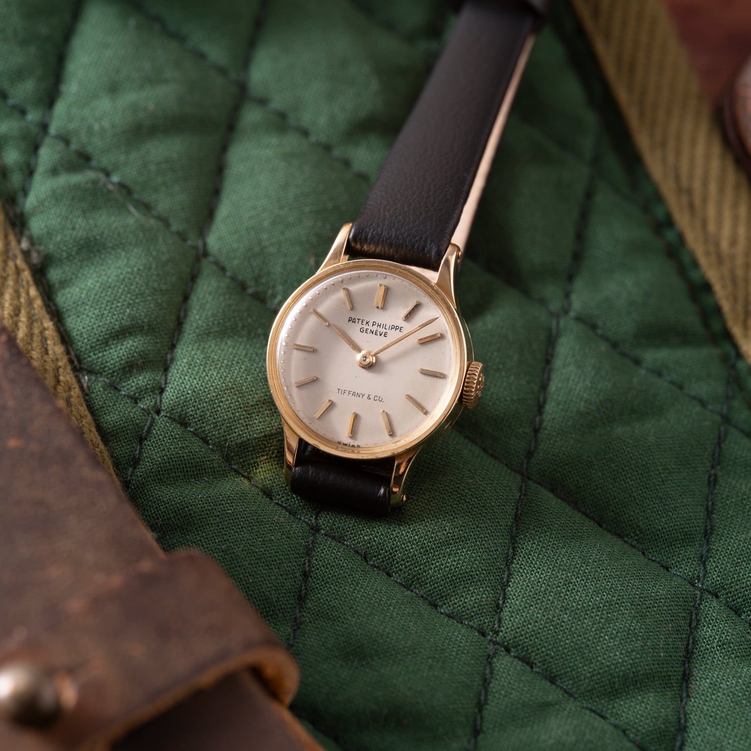 A Patek Philippe Calatrava 'Tiffany & Co' ladies watch with a black leather strap and 18K yellow gold case, displayed on quilted green fabric. The dial features Patek Philippe Genève and Tiffany & Co.