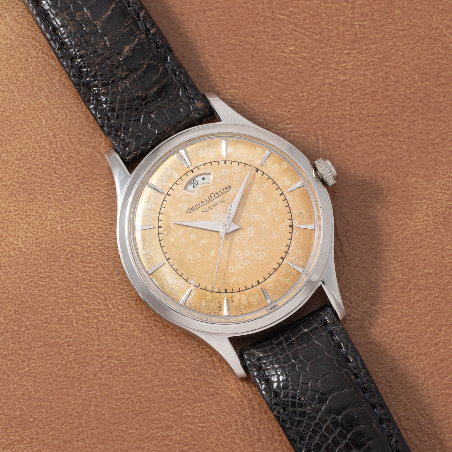 A Jaeger LeCoultre 'European Market' Powermatic wristwatch with a patinated beige dial, silver indices, date window, power-reserve display, and black leather strap is showcased on a brown textured surface.
