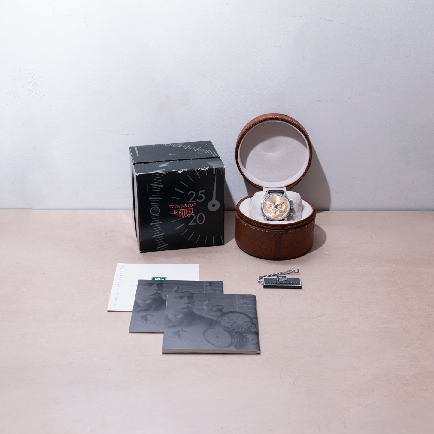 A TAG Heuer 1964 Carrera Re-Edition wristwatch is shown in an open brown case, with its box, informational booklets, tag, and cards arranged on a light surface. The watch box displays aviation-themed graphics.