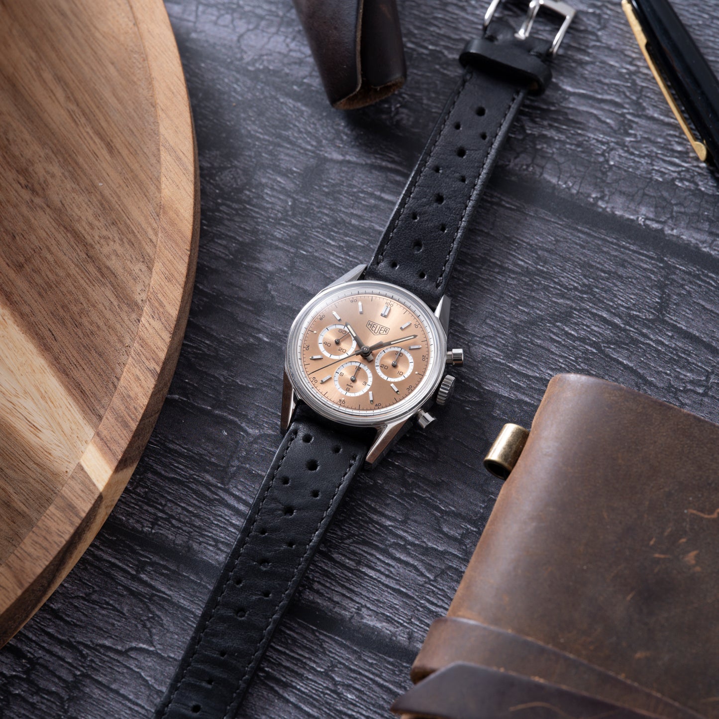 A TAG Heuer 1964 Carrera Re-Edition chronograph with a rose gold dial, black perforated leather strap, and three subdials is shown on a dark wooden surface with a round tray, leather item, and pen nearby.