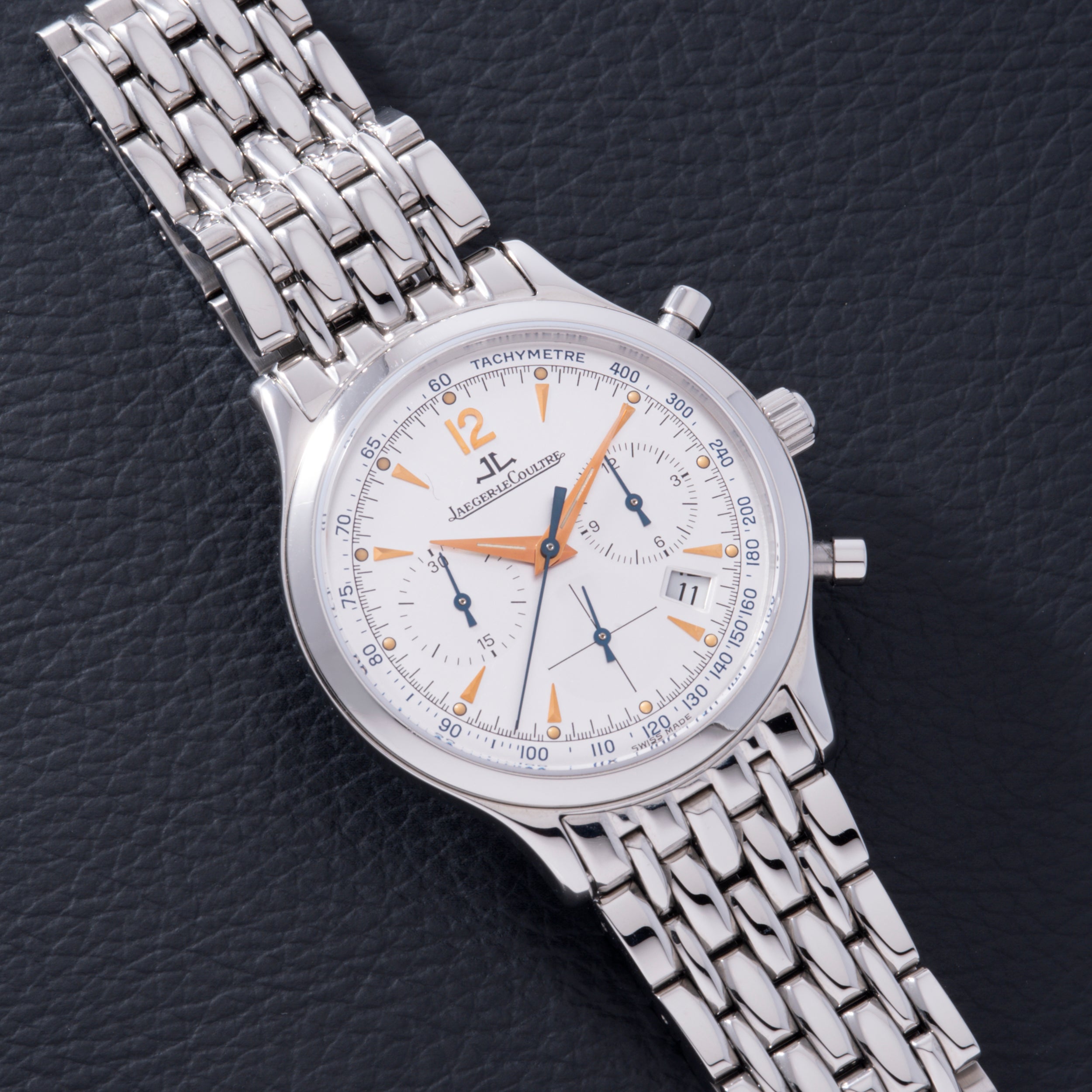 A Jaeger LeCoultre Master Control Chronograph Mecaquartz wristwatch with a white dial, orange accents, and metal link bracelet rests on a black textured surface.