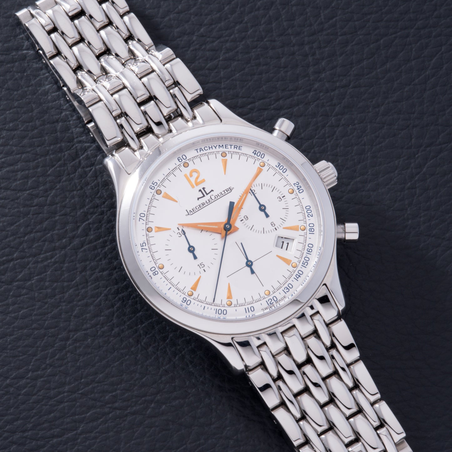 A Jaeger LeCoultre Master Control Chronograph Mecaquartz wristwatch with a white dial, orange accents, and metal link bracelet rests on a black textured surface.