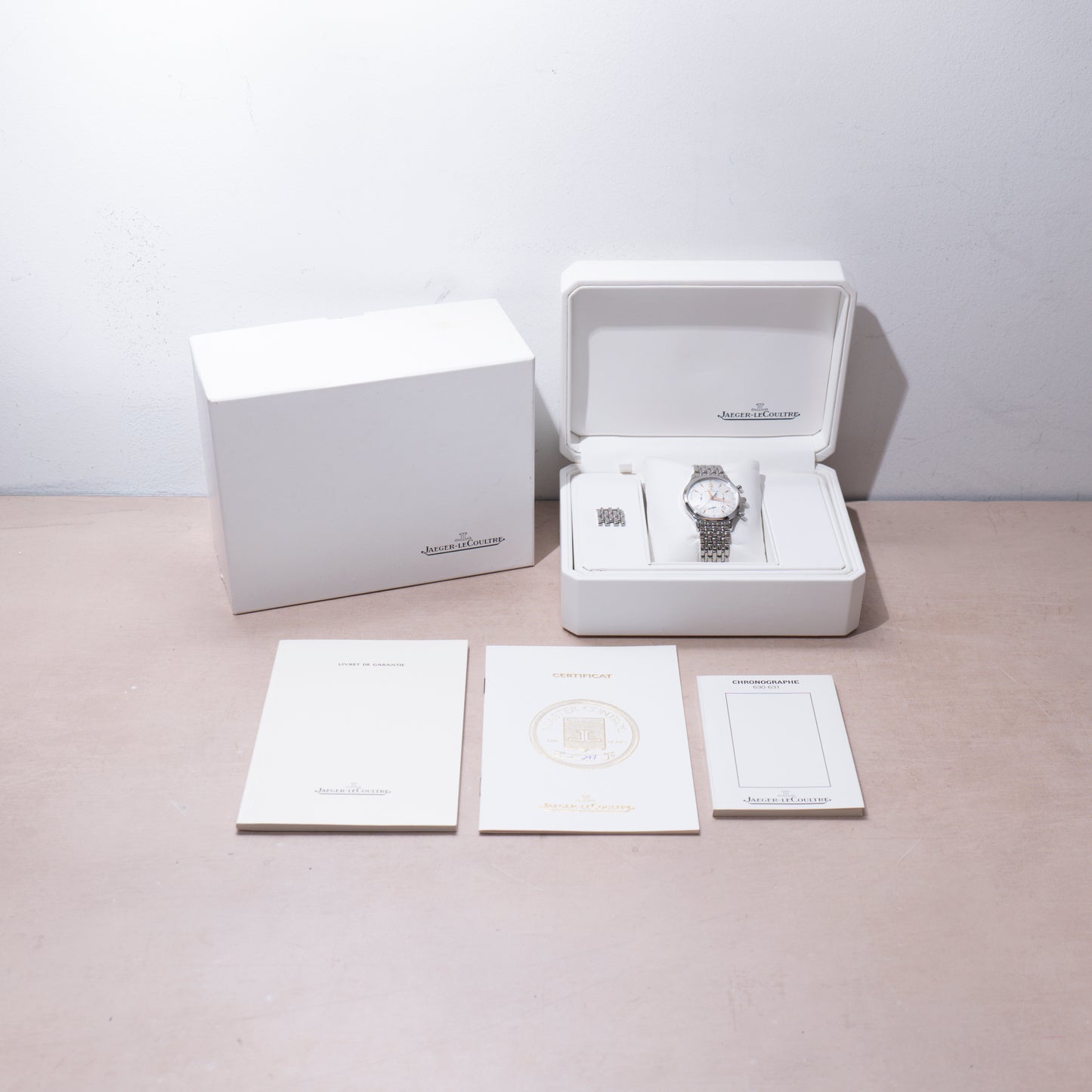 A Jaeger-LeCoultre Master Control Chronograph Mecaquartz wristwatch is displayed in a white box with its outer box, extra link, and three branded white booklets on a beige surface.