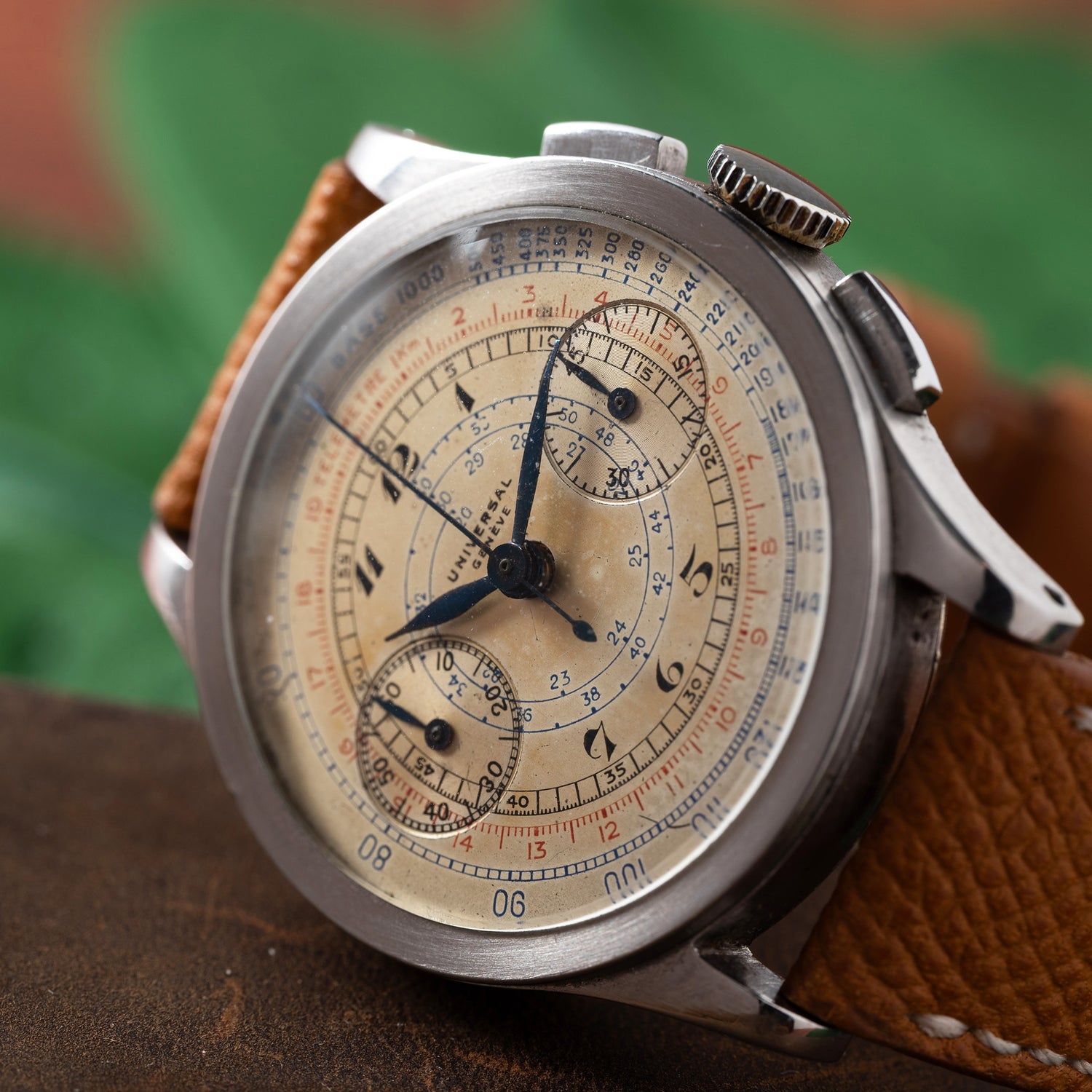 A close-up of the Universal Genève Compax 'Breguet' chronograph wristwatch by Universal Geneve, featuring a beige dial with intricate markings, three subdials, a brown leather strap, and a silver case against a blurred green and brown background.