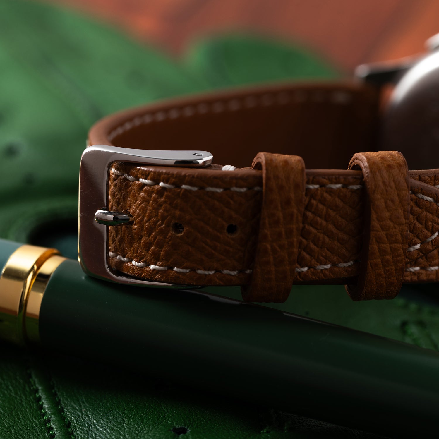 A close-up of a brown leather strap with white stitching sits beside a dark green fountain pen—an ideal pairing for your Universal Genève Compax 'Breguet' by Universal Geneve.
