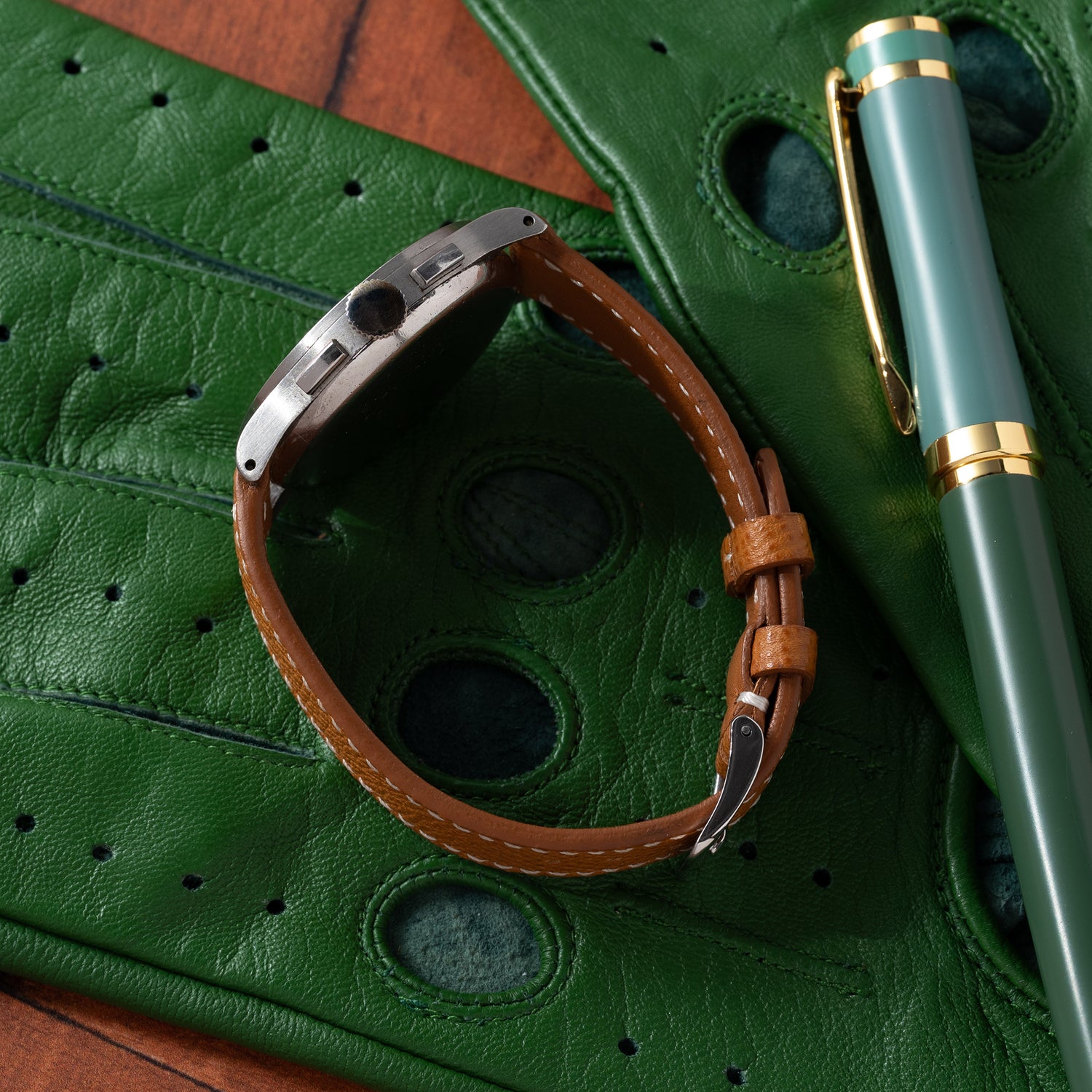 A Universal Genève Compax 'Breguet' chronograph with a brown leather strap rests on green leather gloves, beside a green and gold fountain pen on a wooden surface.