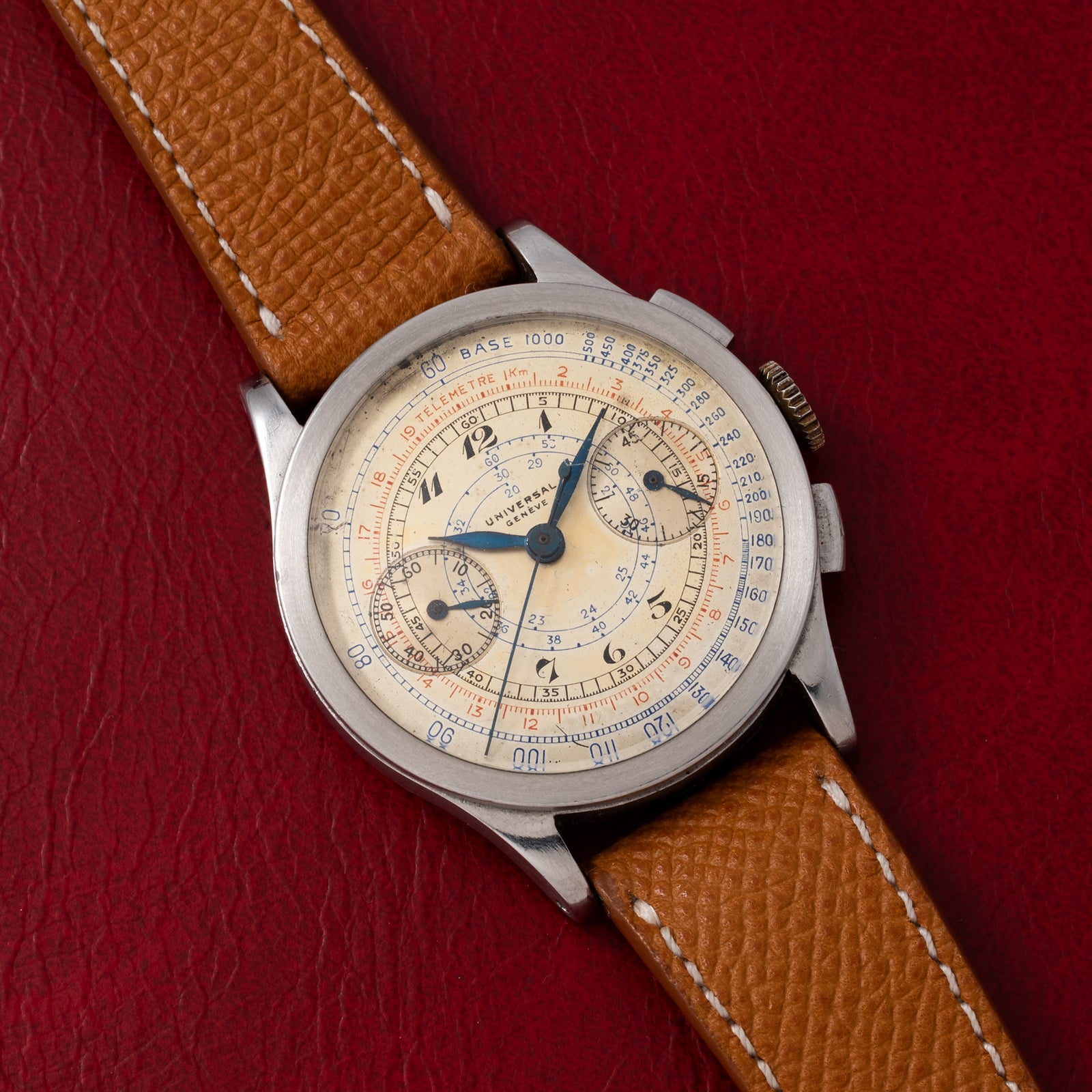 The Universal Genève Compax 'Breguet' by Universal Geneve, a vintage chronograph with a brown leather strap, stainless steel case, and cream dial with blue, red, and black subdials, rests on textured red leather.