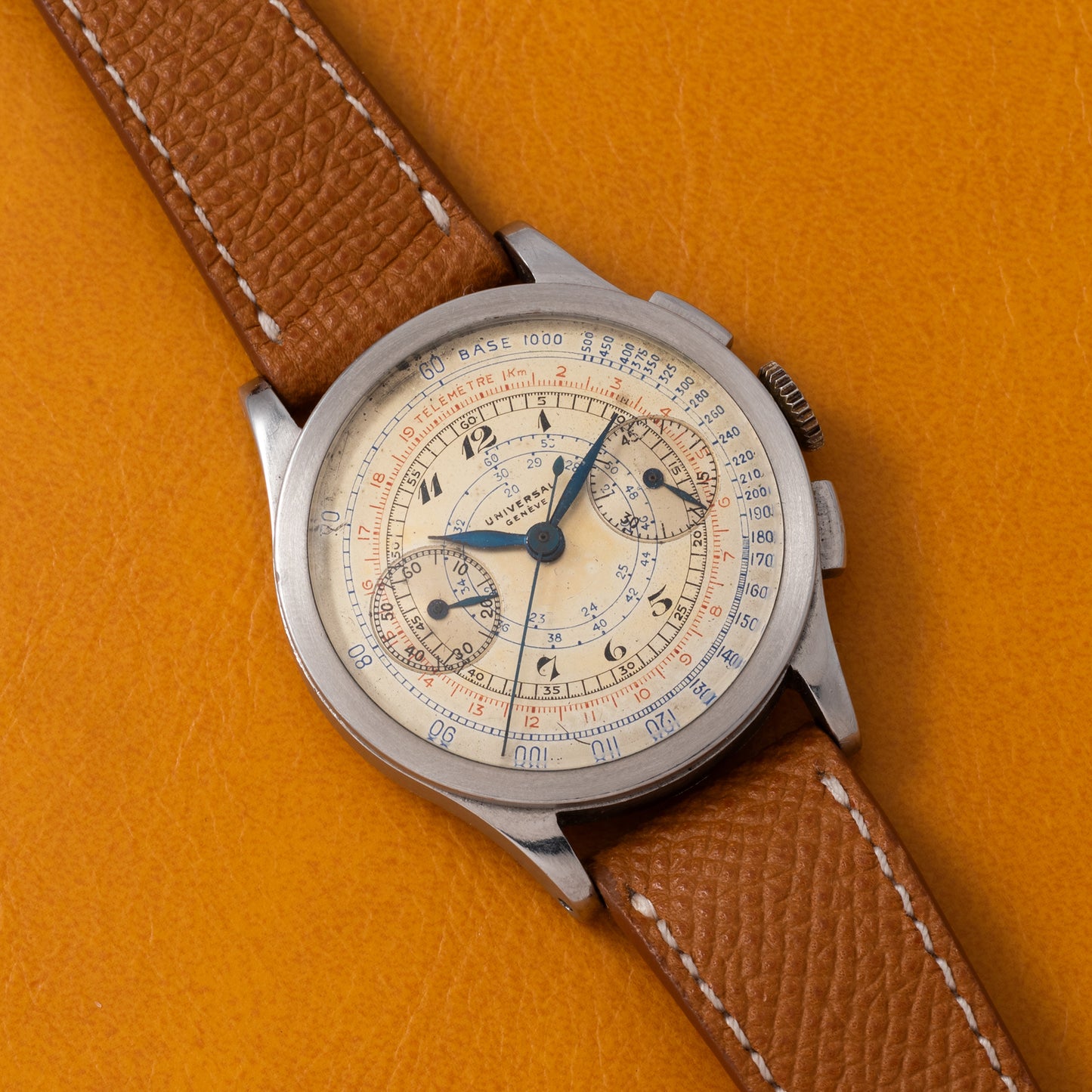 The Universal Geneve Compax 'Breguet' wristwatch, featuring a brown leather strap and a detailed cream dial with subdials and scales, is displayed on an orange textured surface.