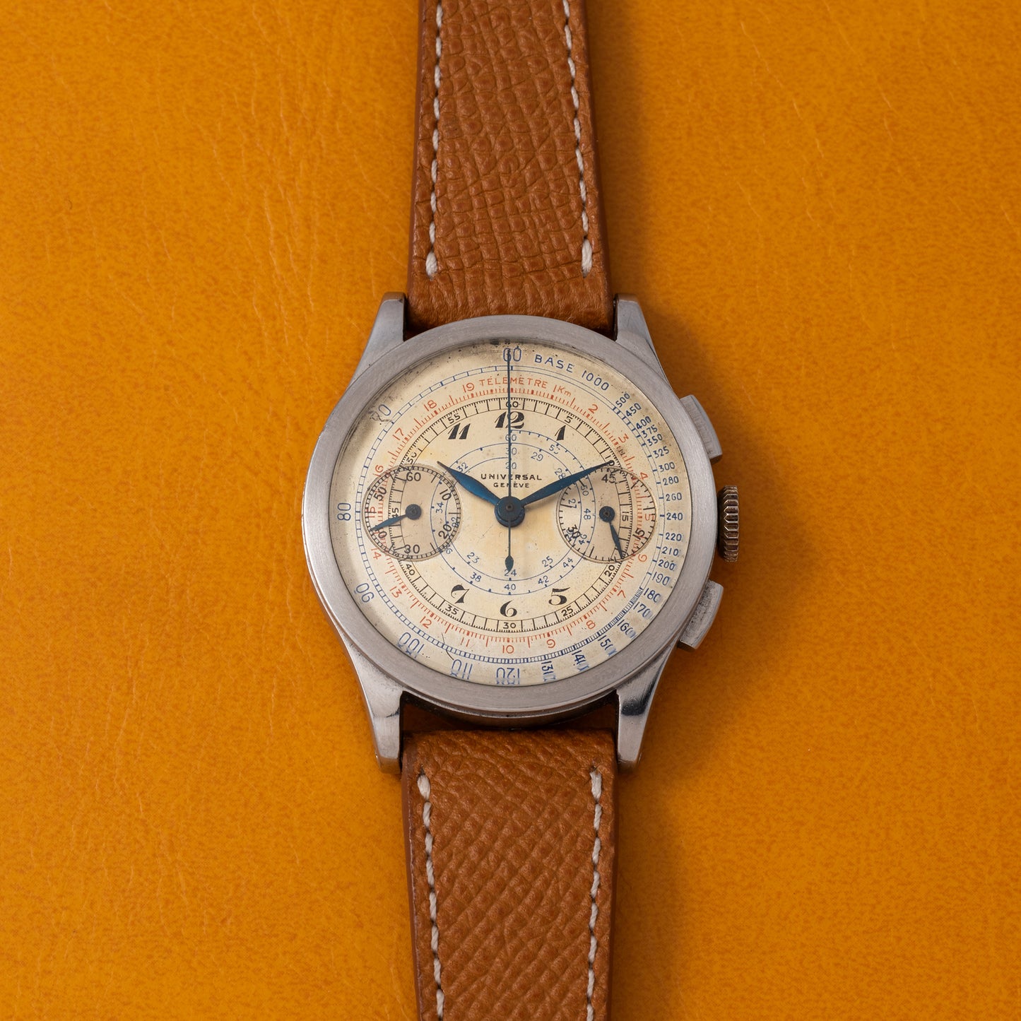 A vintage Universal Genève Compax 'Breguet' chronograph with a brown leather strap and a cream dial with blue hands and multiple subdials, displayed on an orange textured surface.