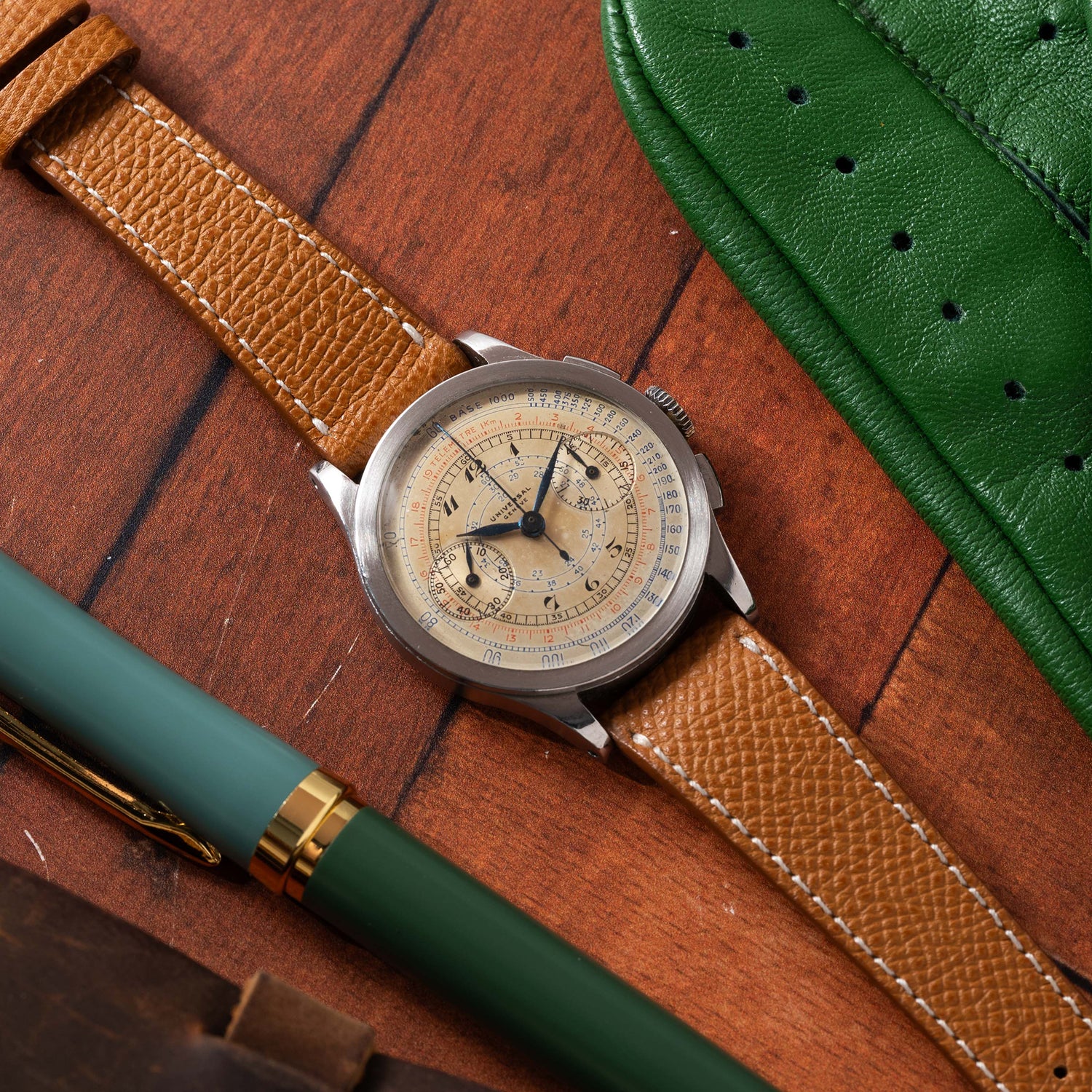 A Universal Genève Compax 'Breguet' vintage chronograph by Universal Geneve with a tan leather strap and cream dial rests on a wooden surface beside a green pen and green leather case.