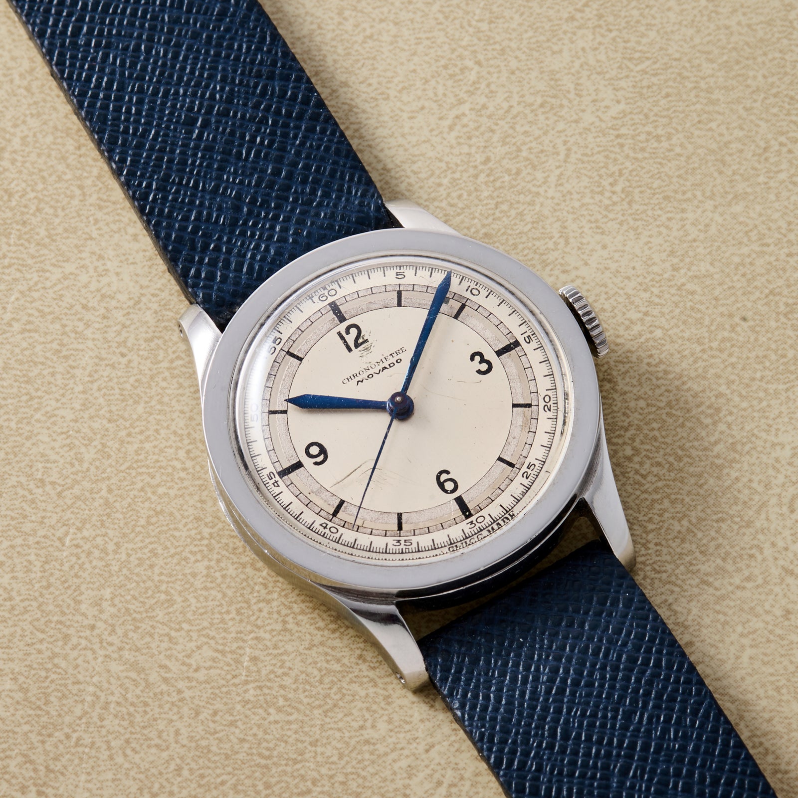 The Movado 'Calatrava' Steel, featuring a cream dial, blue hands, bold black numerals, and a textured dark blue leather strap, displays Swiss elegance as it's showcased on a beige surface.