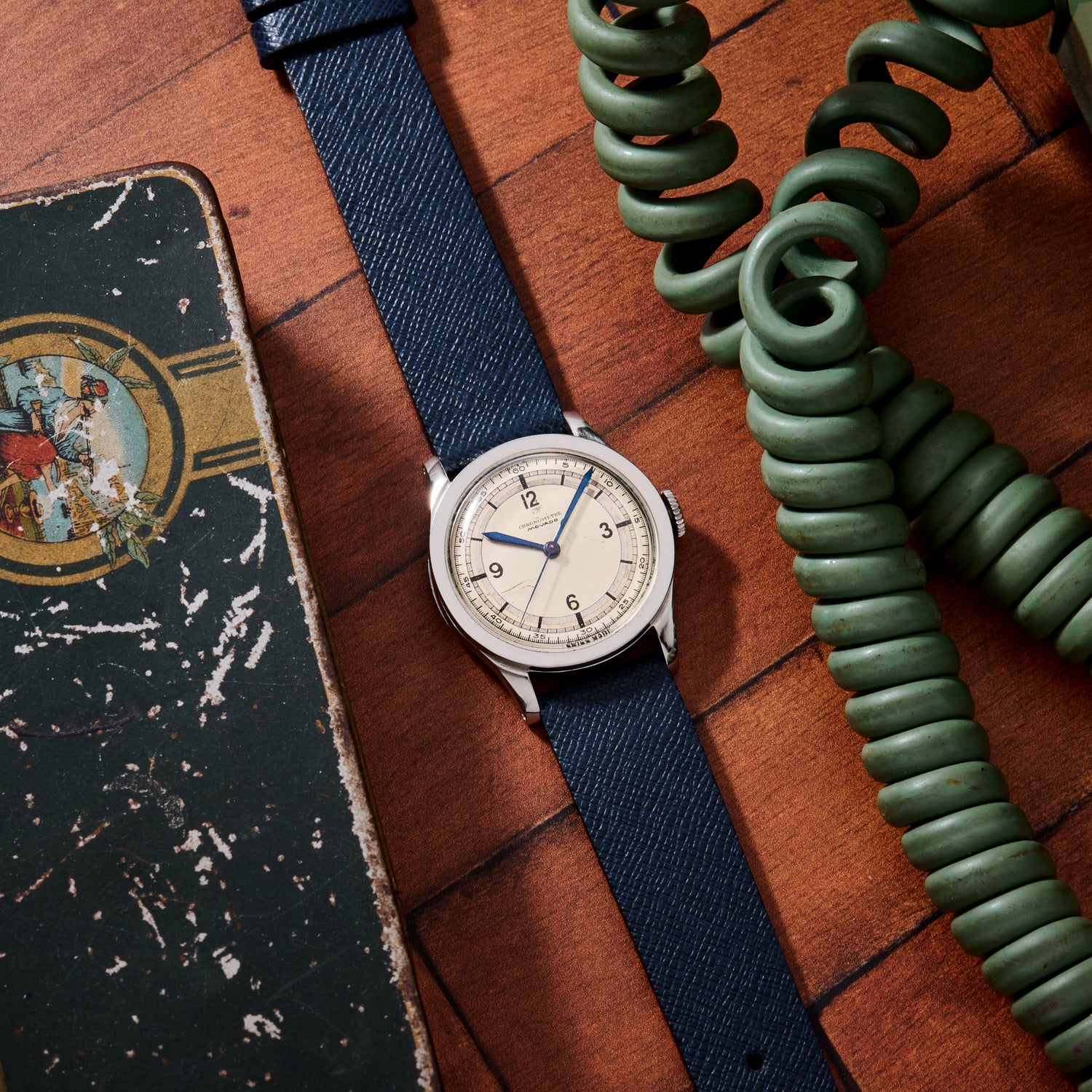 The Movado 'Calatrava' Steel watch with a navy strap rests on a wooden surface beside a green coiled phone cord and a worn illustrated box, capturing the timeless charm of Swiss watchmaking.