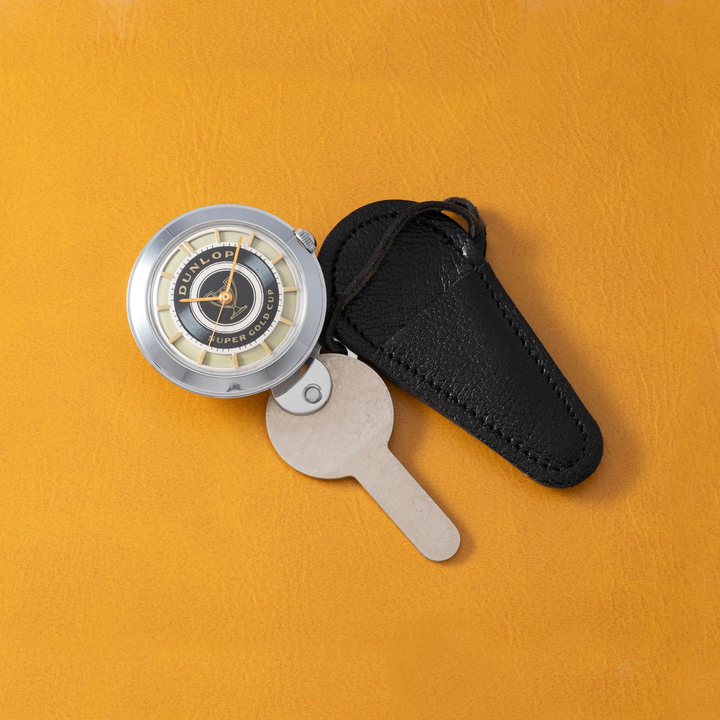 A vintage Prestige Watch Co. Dunlop Key Watch, featuring a round silver and black design on a beige key-shaped tag with a black leather case—perfect for classic car fans—rests on an orange leather surface.