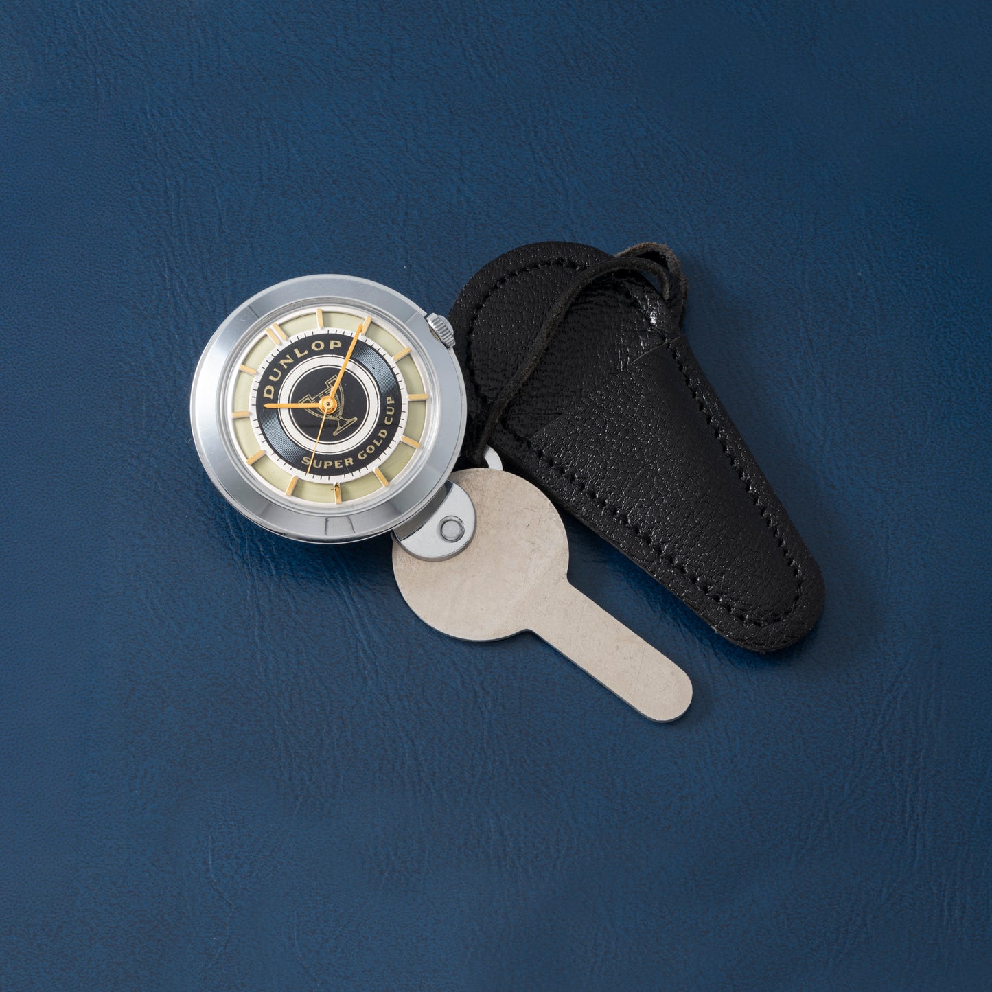 A vintage Dunlop Prestige Watch Co. key watch with a metal key and black leather case is perfect for collectors, displayed on a blue textured surface.