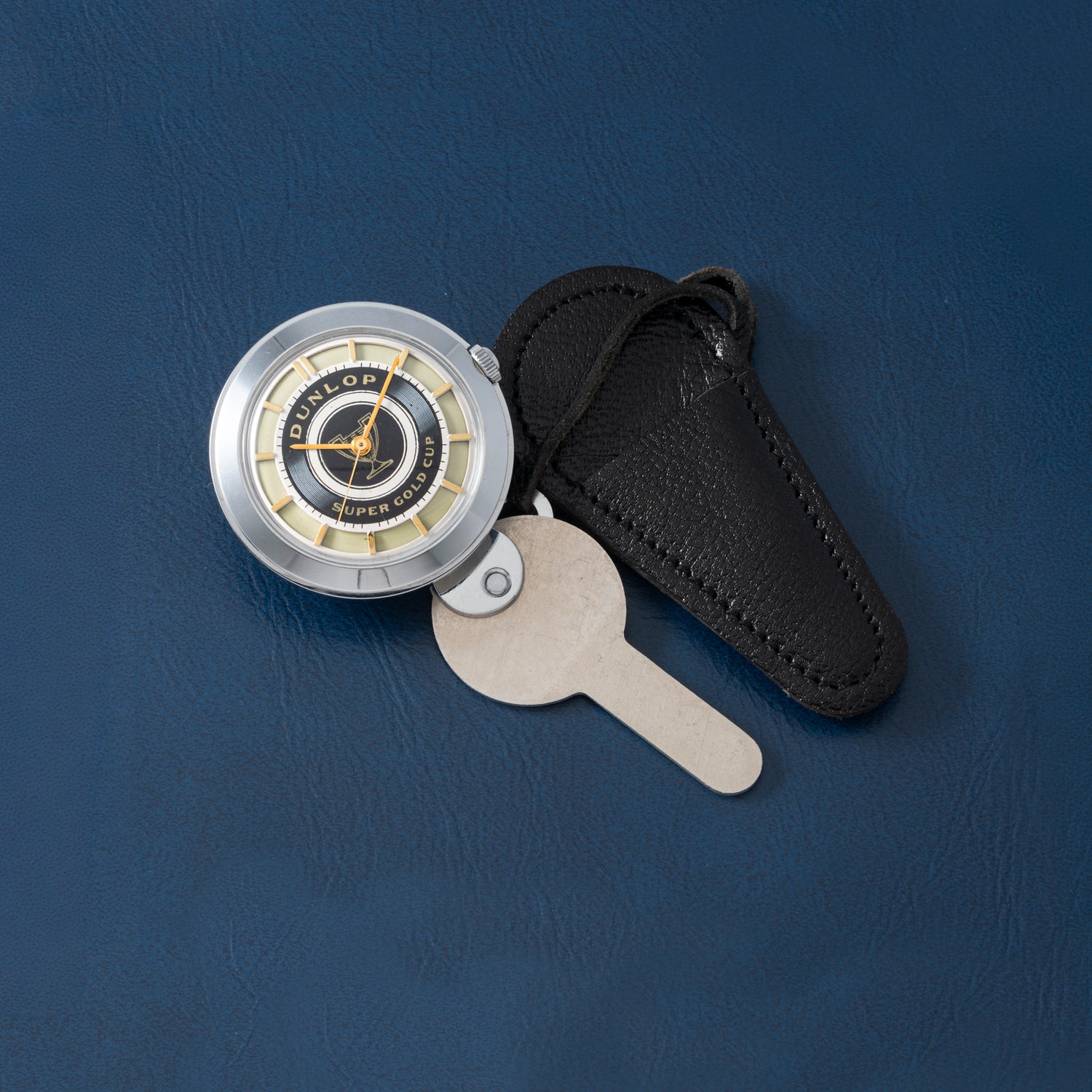 A vintage Dunlop Prestige Watch Co. key watch with a metal key and black leather case is perfect for collectors, displayed on a blue textured surface.