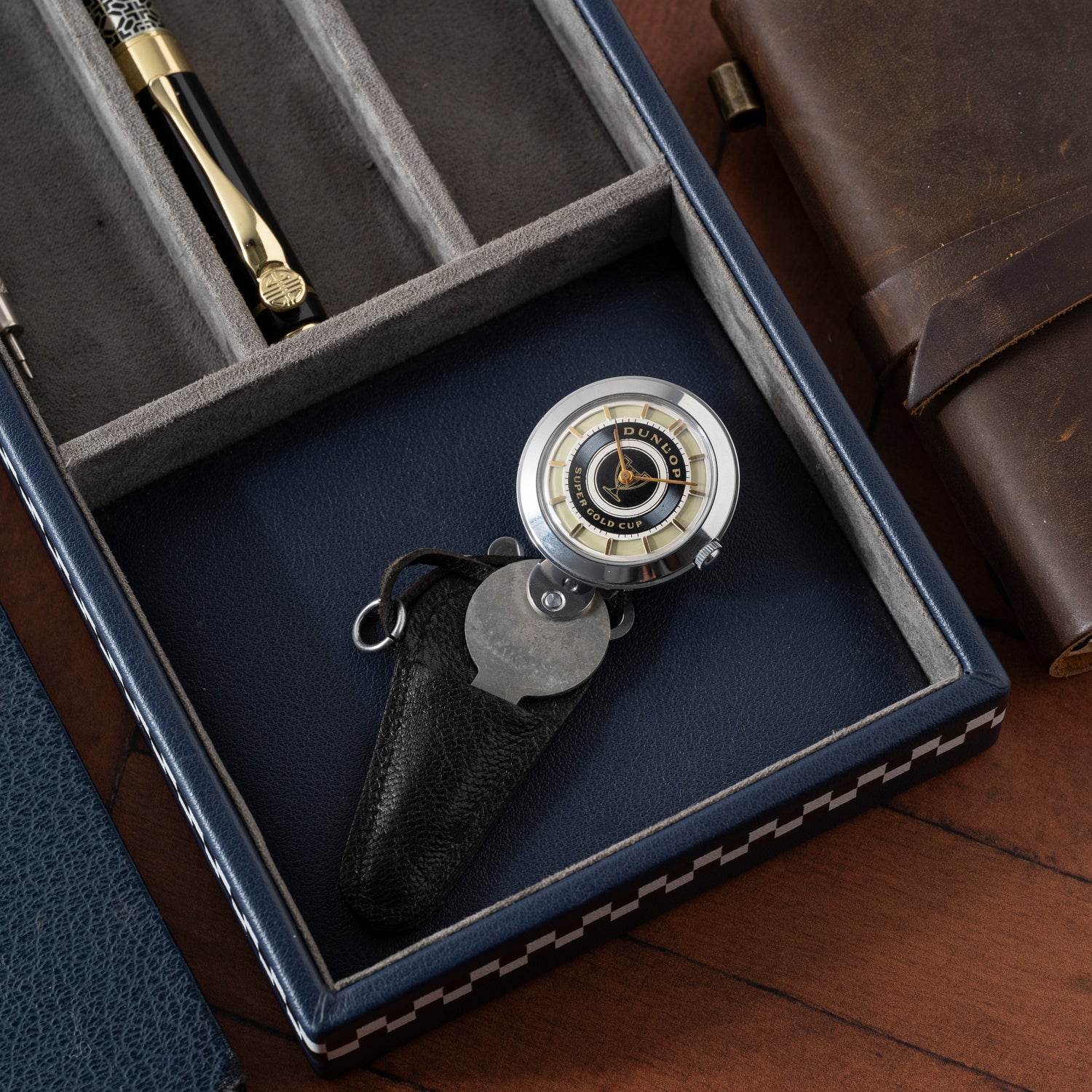A close-up of the Dunlop Prestige Watch Co. Dunlop Key Watch beside a brown leather journal in a blue and grey case—a perfect gift for any classic car enthusiast.