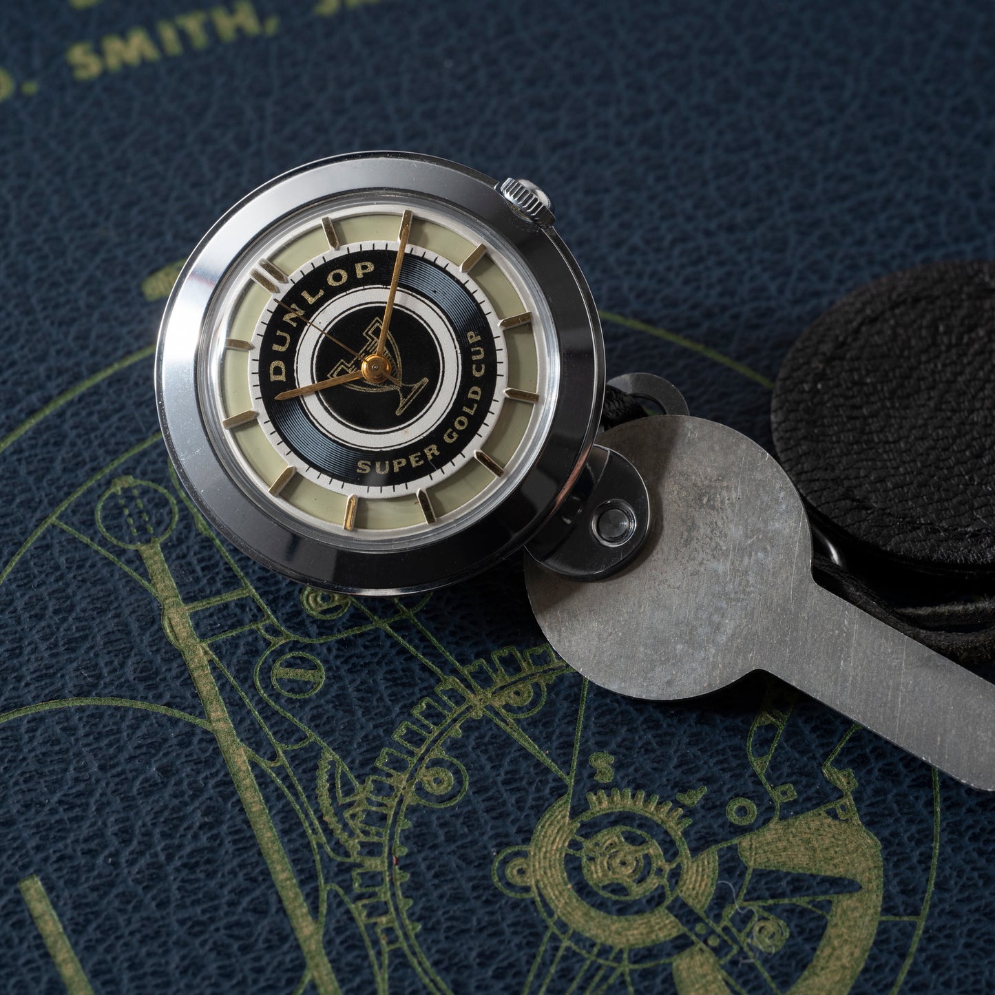 A round Dunlop Prestige Watch Co. Dunlop Key Watch with a black and gold face rests beside a metal key on a textured blue surface featuring yellow technical illustrations—an ideal accessory for classic car enthusiasts.