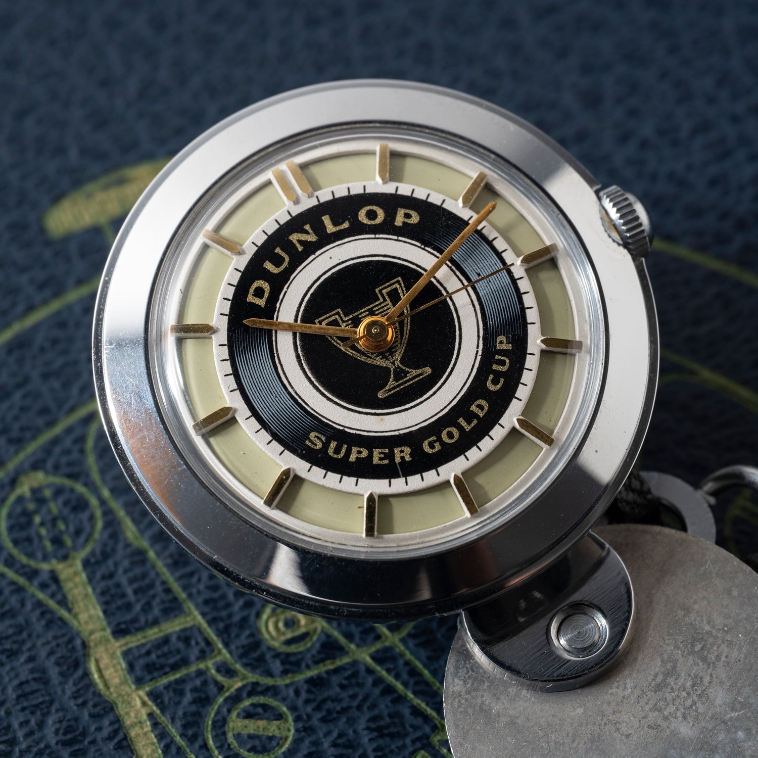A close-up of the Dunlop Prestige Watch Co. Dunlop Key Watch, featuring a black and gold center inscribed with "Dunlop Super Gold Cup," gold hands, and a silver case, set on a textured dark blue surface with subtle yellow accents.