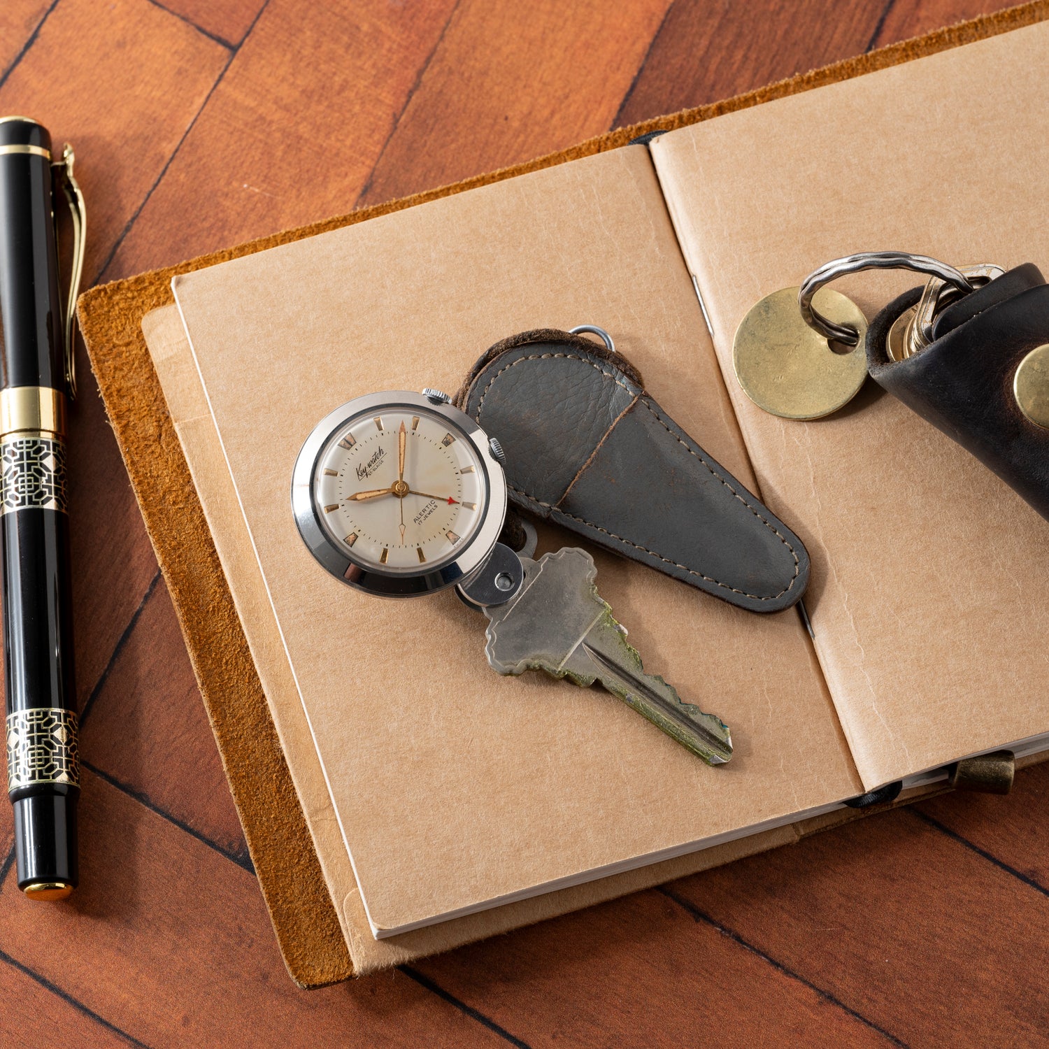 The Saint Blaise Key Watch Alarm, a leather key holder with keys, and a black and gold pen rest on an open brown notebook atop a wooden surface—an ideal set for the classic car enthusiast.