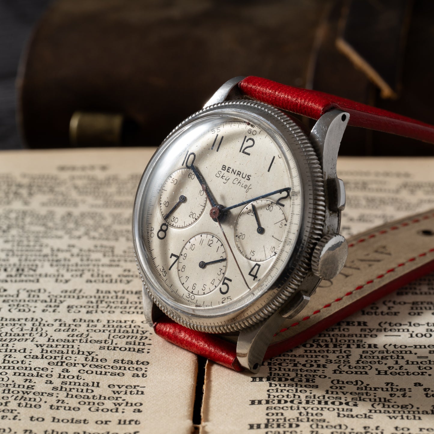 The Benrus Sky Chief by Benrus, featuring a red leather strap, rests on an open, aged book. Its white face sports three subdials and black numerals, set against a softly blurred background that highlights its vintage charm.