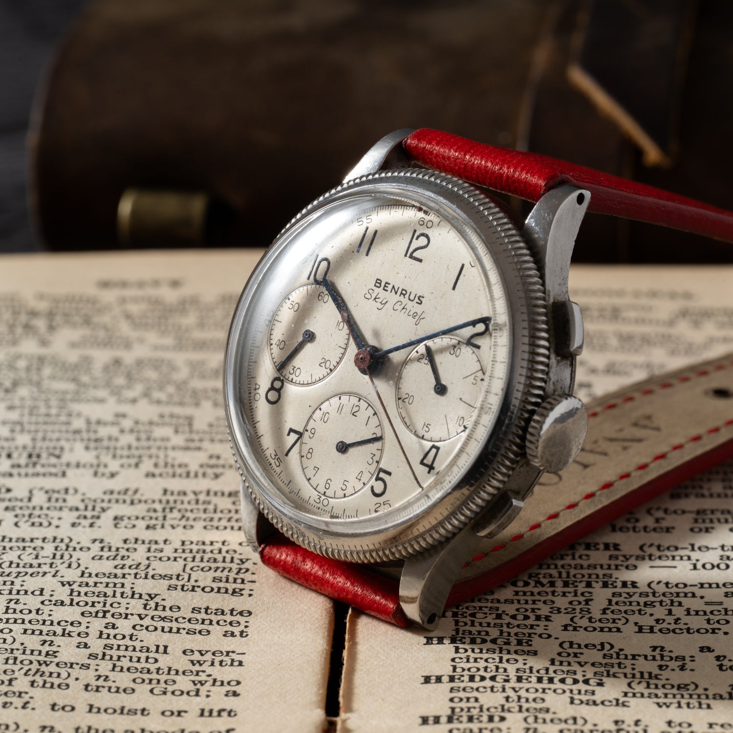 The Benrus Sky Chief by Benrus, featuring a red leather strap, rests on an open, aged book. Its white face sports three subdials and black numerals, set against a softly blurred background that highlights its vintage charm.