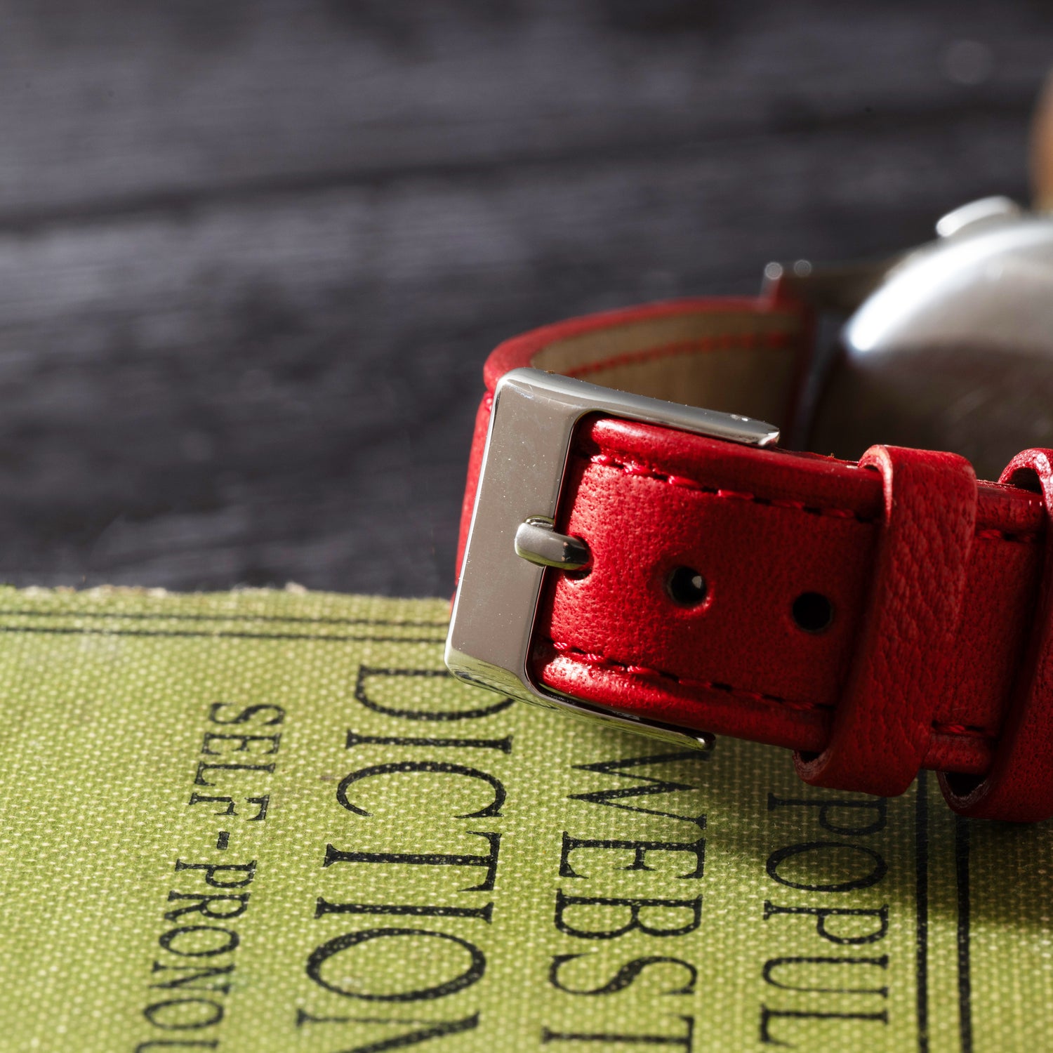 A close-up of the Benrus Sky Chief’s red leather strap with a silver buckle, evoking vintage pilot style, rests on a green Websters Dictionary atop a dark wooden surface.