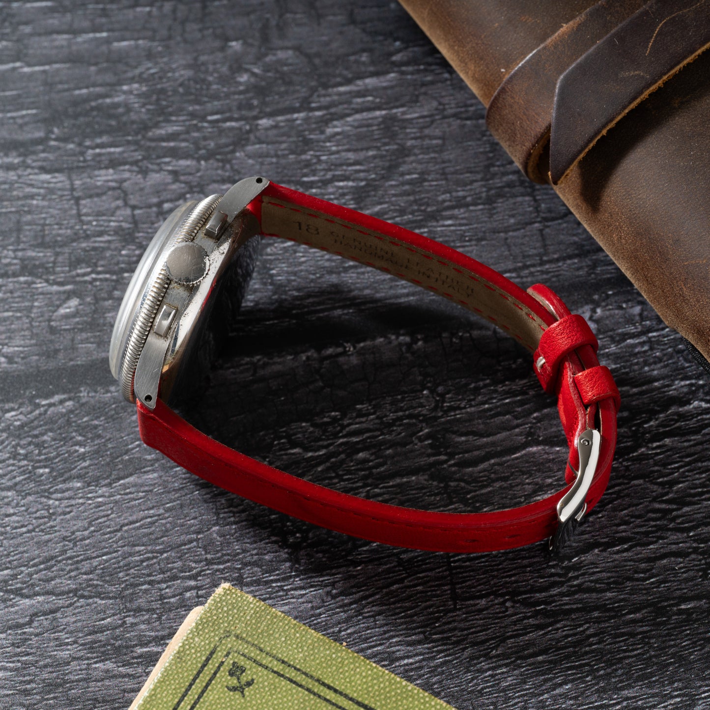 The Benrus Sky Chief, a vintage pilot watch with a silver case and red leather strap, rests on a dark textured surface near a brown leather item and the corner of a green book.