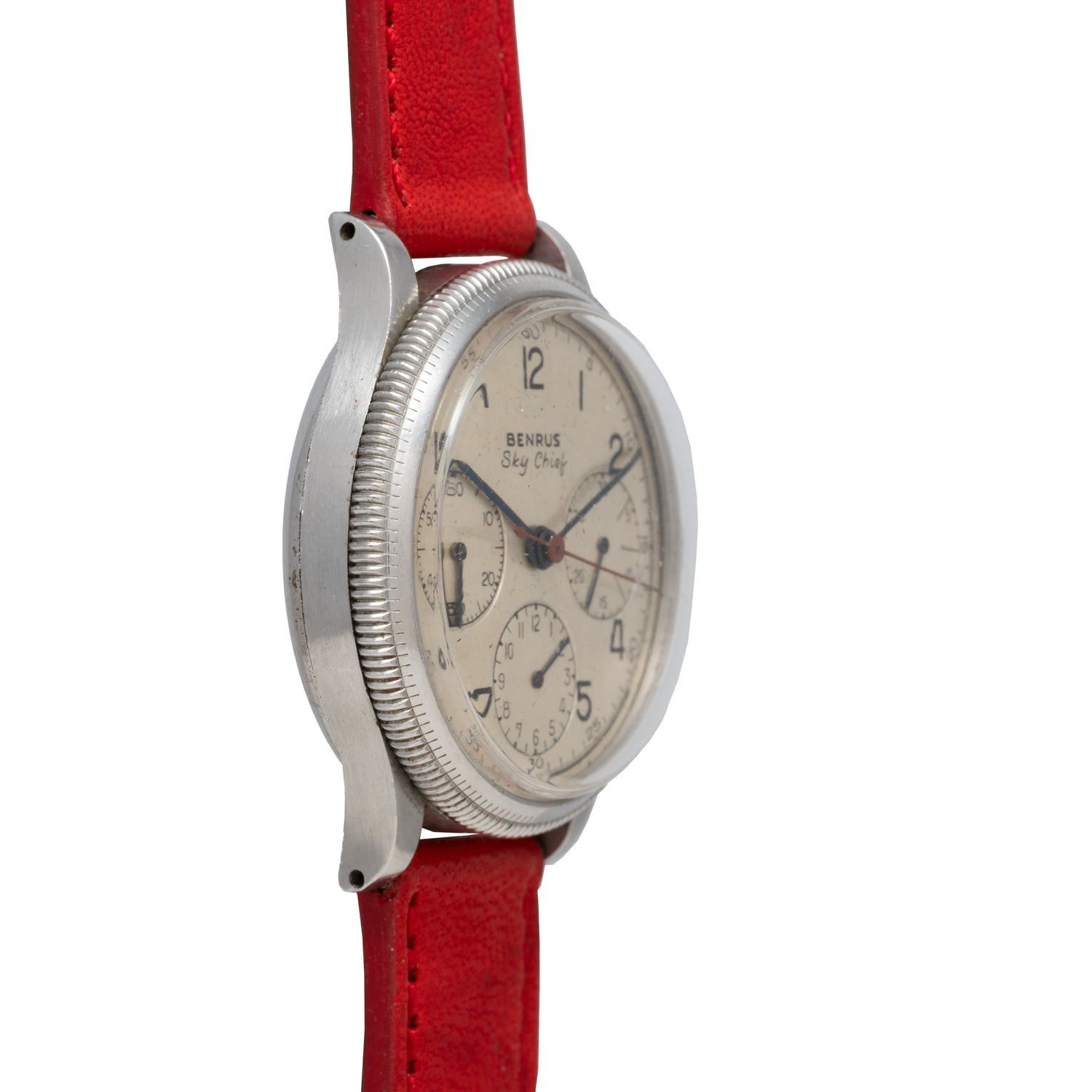 The Benrus Sky Chief by Benrus is a vintage pilot watch with a round silver case, cream dial with black numerals and subdials, displayed at an angle on a red leather strap against a white background.