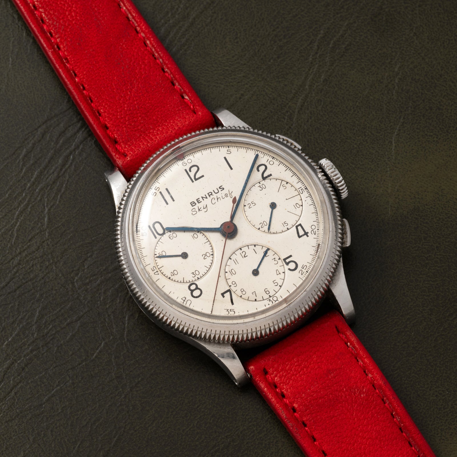 A vintage Benrus Sky Chief by Benrus, featuring a white dial with three subdials, silver case, and red leather strap, is shown on a dark green textured surface.