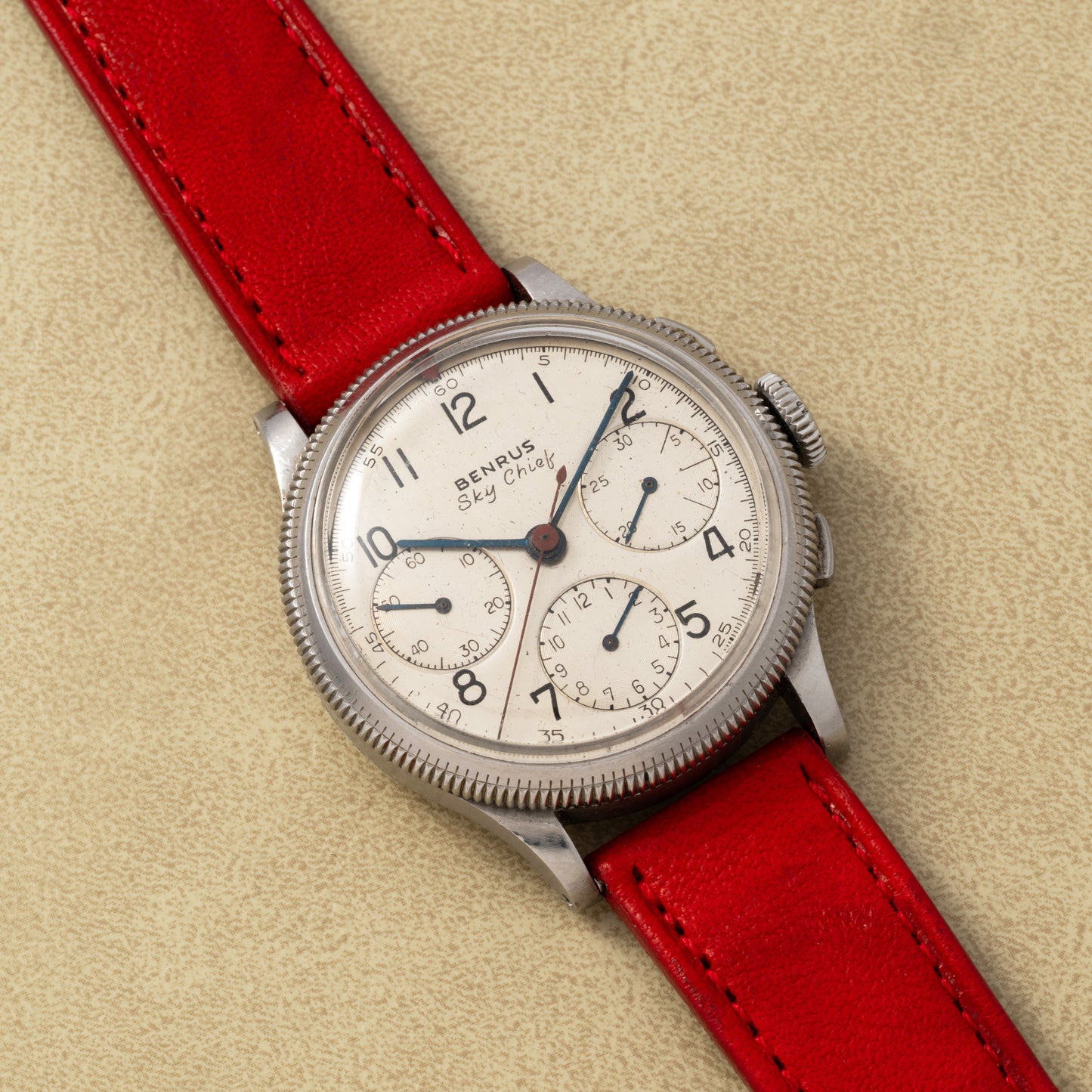The Benrus Sky Chief by Benrus is a vintage pilot chronograph with a cream dial, black numerals, three subdials, and a red leather strap, displayed on a light tan surface.