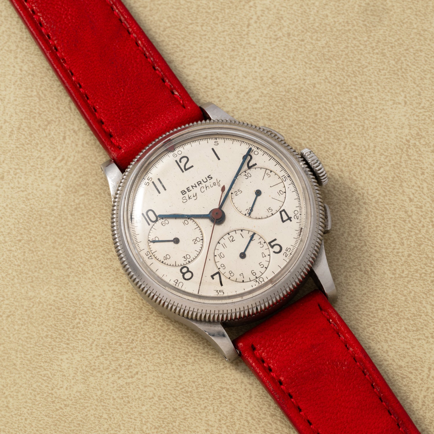 The Benrus Sky Chief by Benrus is a vintage pilot chronograph with a cream dial, black numerals, three subdials, and a red leather strap, displayed on a light tan surface.