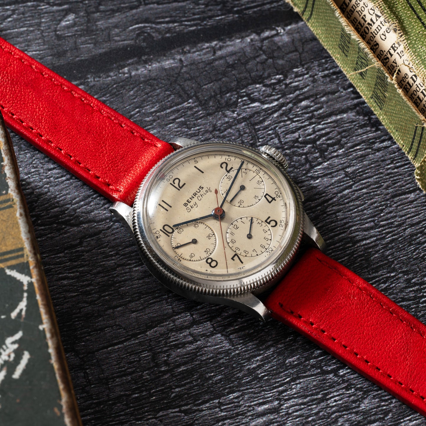 The Benrus Sky Chief, a vintage pilot chronograph by Benrus, features a beige dial with black numerals and subdials, paired with a red leather strap. It rests on a dark textured surface beside scattered papers and books.