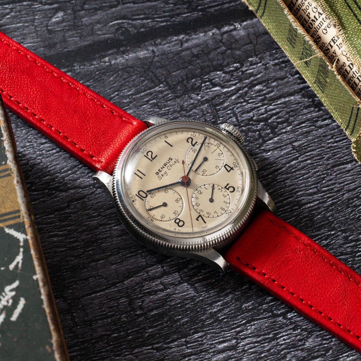 The Benrus Sky Chief, a vintage pilot chronograph by Benrus, features a beige dial with black numerals and subdials, paired with a red leather strap. It rests on a dark textured surface beside scattered papers and books.