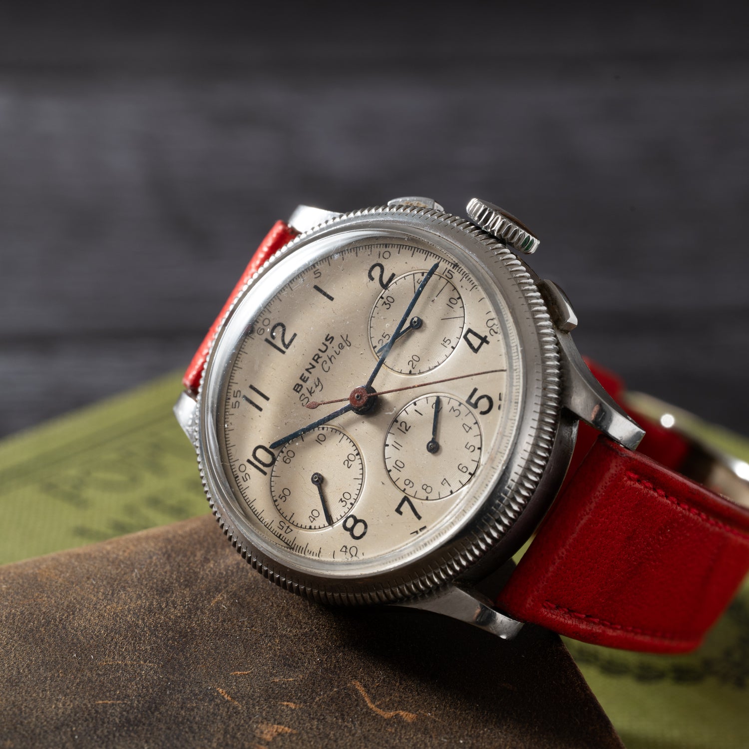 A Benrus Sky Chief pilot chronograph with a cream dial, black numerals, and three subdials sits on a closed book. It features a bright red leather strap and is slightly angled to the left. Brand: Benrus. Product Name: Benrus Sky Chief.