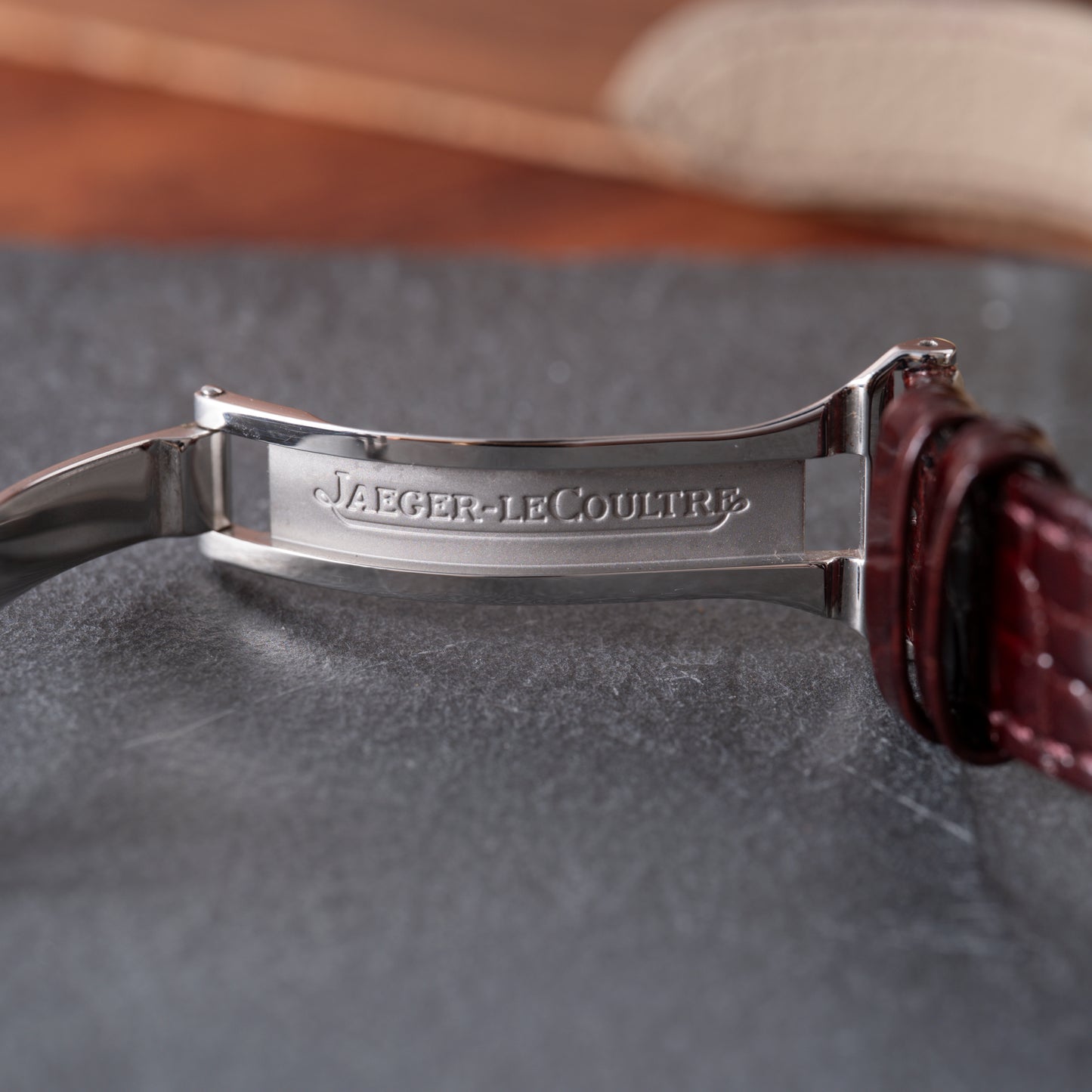 A close-up of the Jaeger LeCoultre Master Control Perpetual Calendar clasp, engraved with the brand name, attached to a brown leather strap on a gray textured surface.