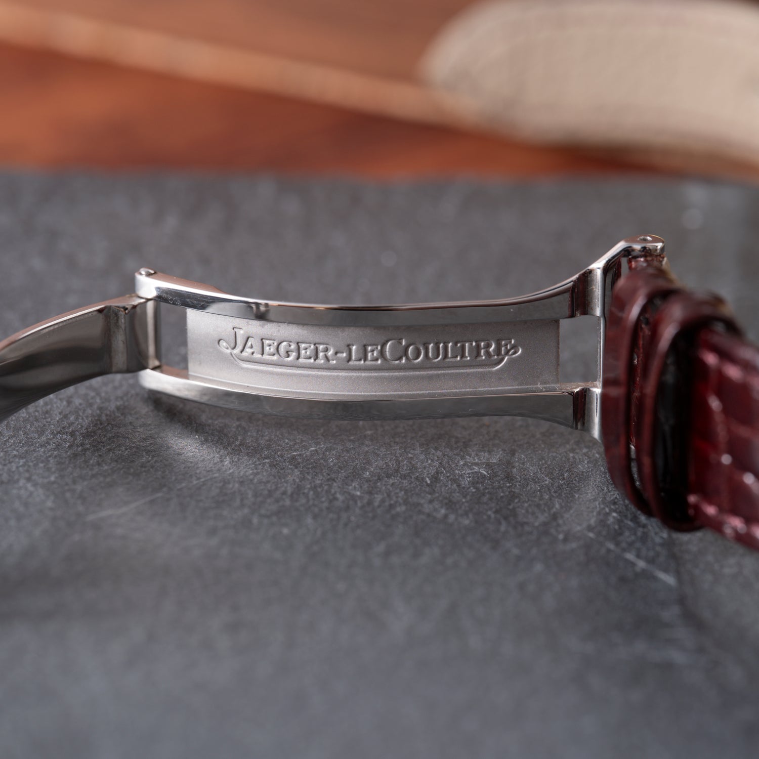 A close-up of the Jaeger LeCoultre Master Control Perpetual Calendar clasp, engraved with the brand name, attached to a brown leather strap on a gray textured surface.