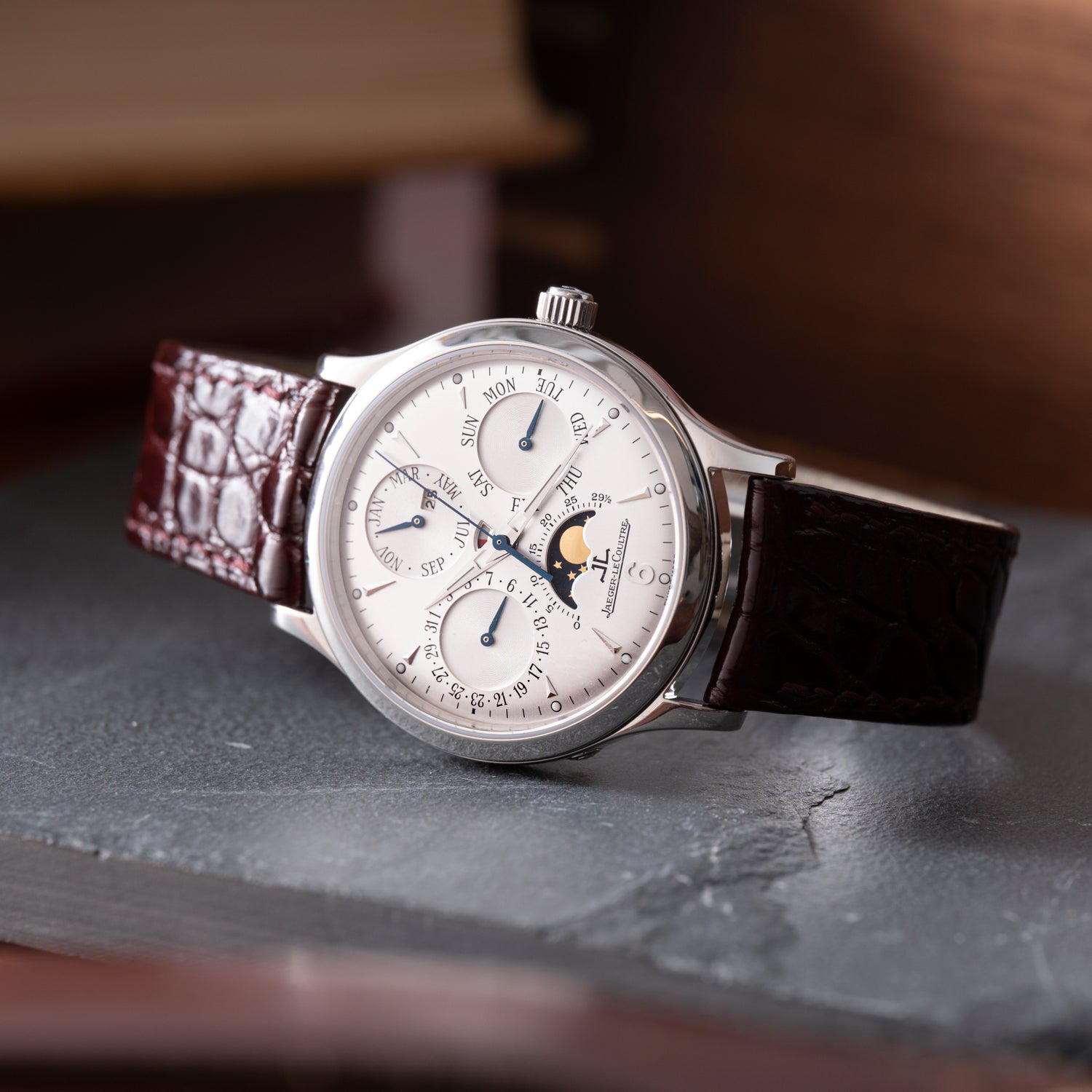 The Jaeger LeCoultre Master Control Perpetual Calendar is a vintage watch with a silver case, white dial featuring subdials and moon phase, and a brown crocodile-pattern leather strap displayed on a gray surface.