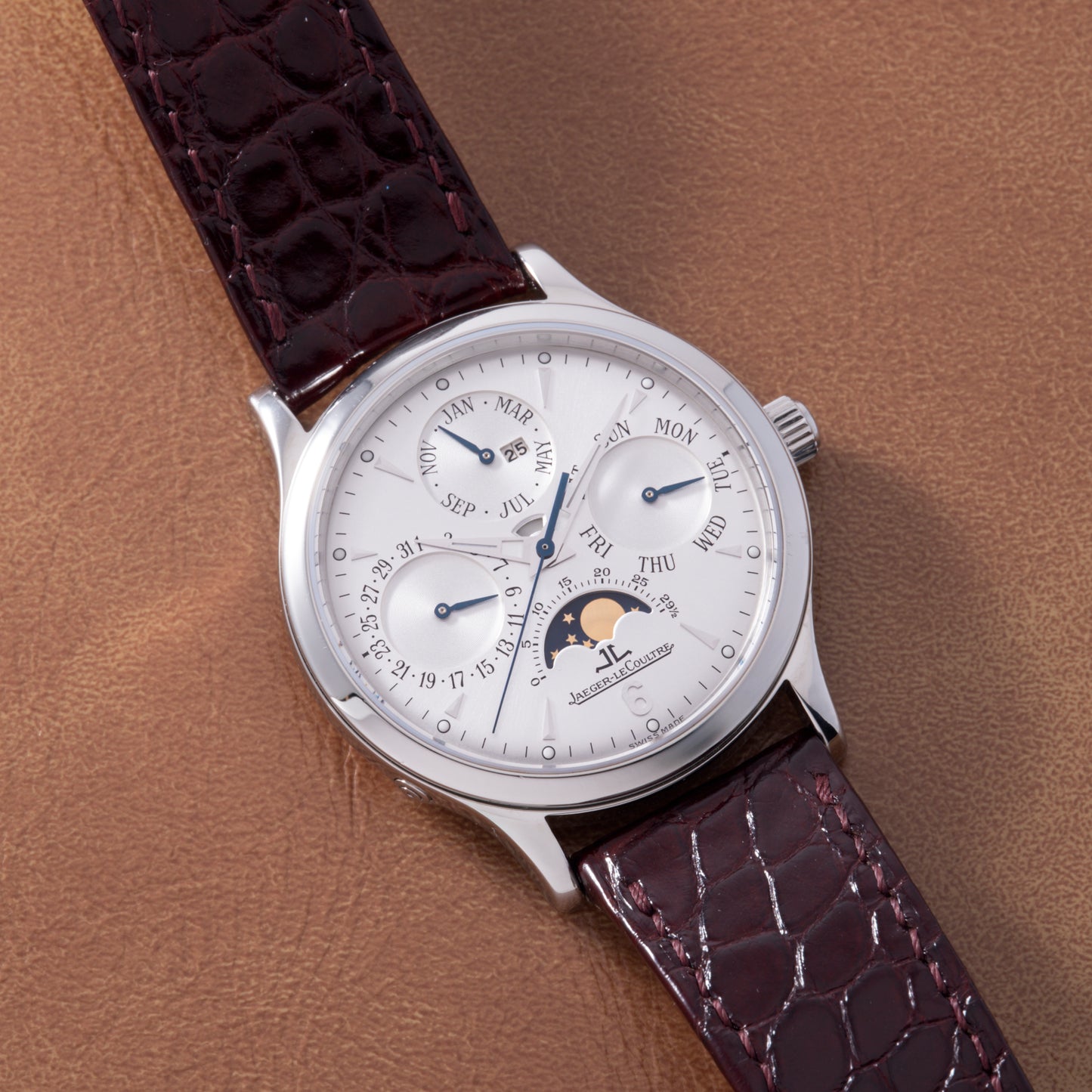 A Jaeger LeCoultre Master Control Perpetual Calendar wristwatch with a silver dial, moonphase, and calendar subdials sits on a brown leather surface, complemented by its brown leather strap.