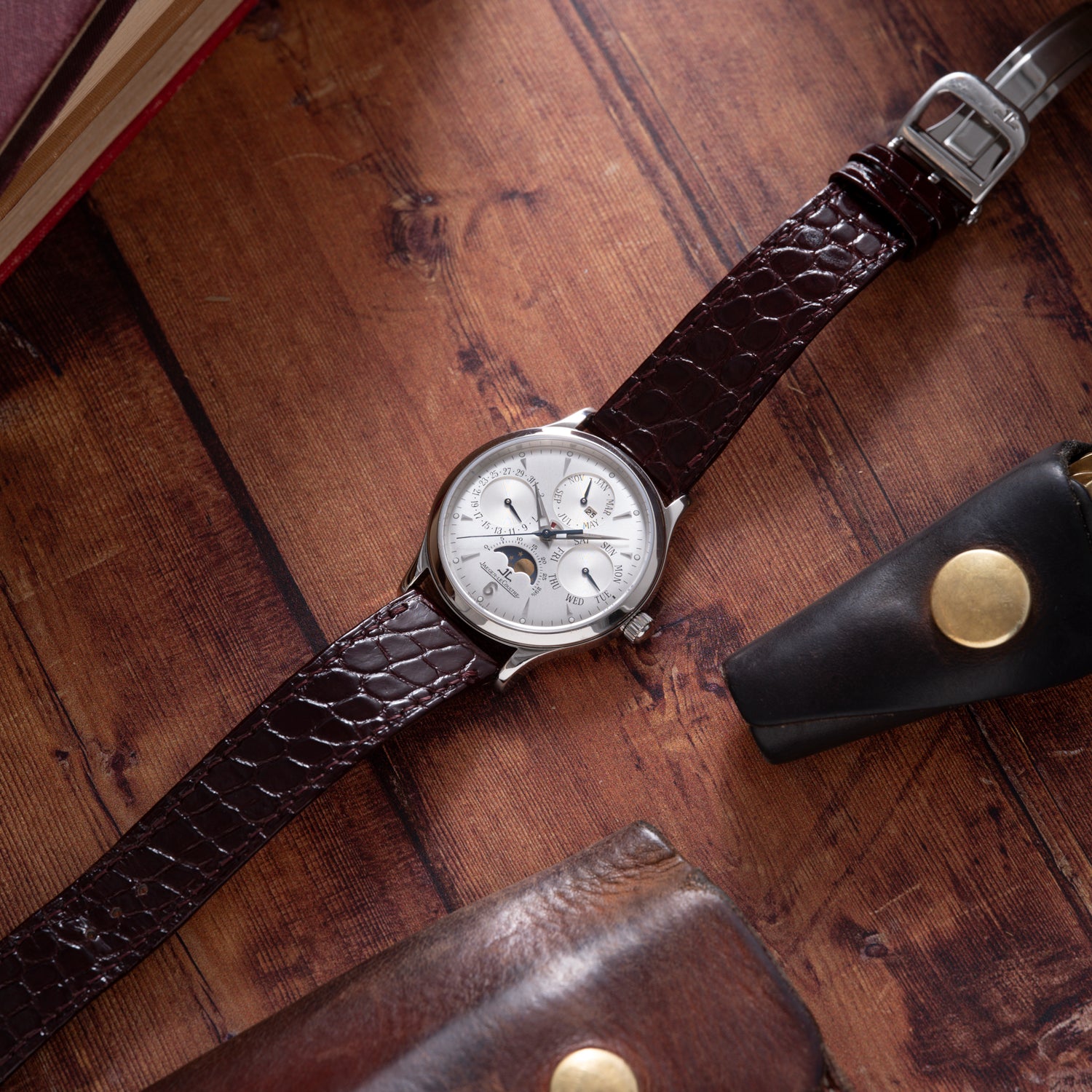 The Jaeger-LeCoultre Master Control Perpetual Calendar, featuring a silver dial with multiple subdials and a brown leather strap, rests on a wooden surface beside a leather case and book.