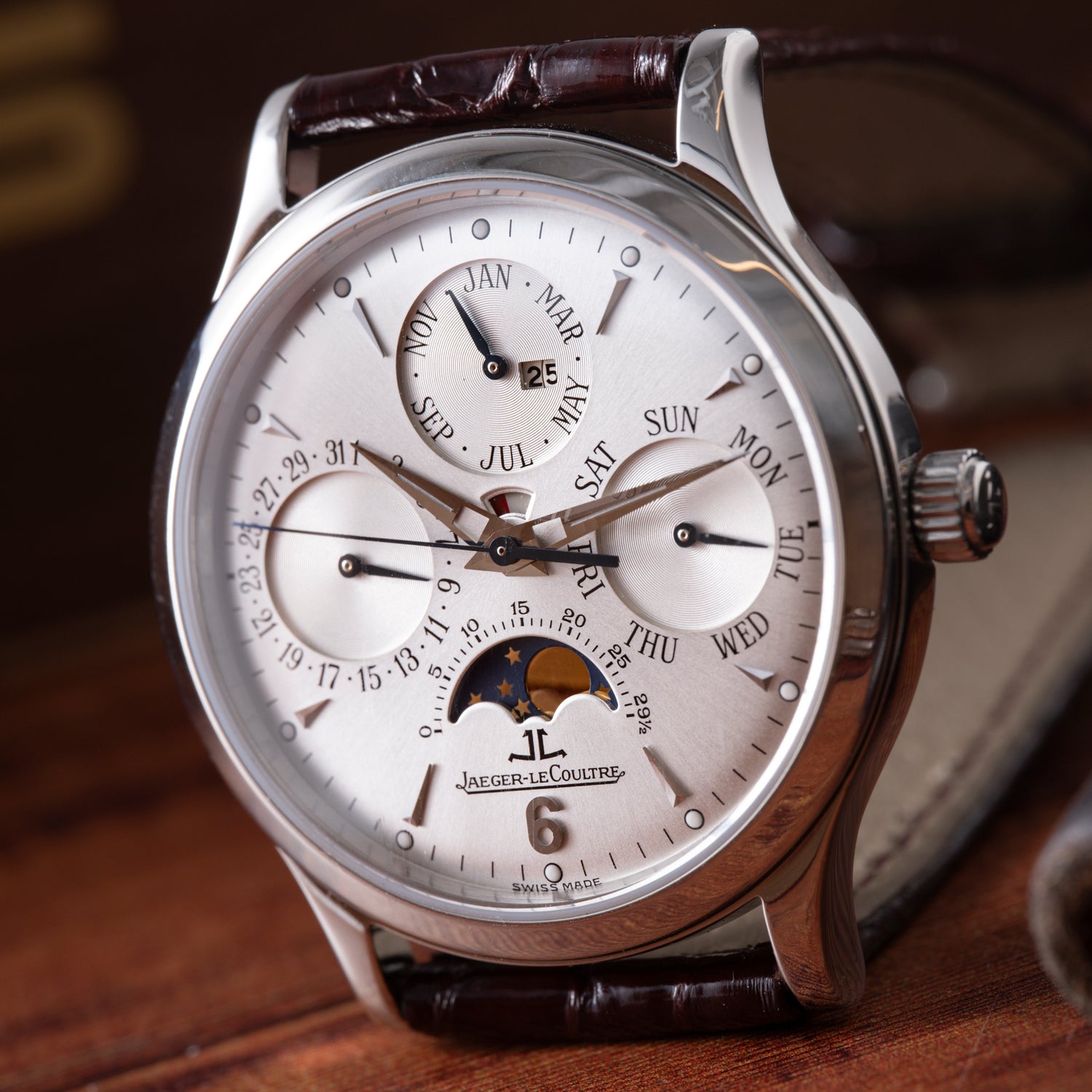 A close-up of the Jaeger-LeCoultre Master Control Perpetual Calendar wristwatch by Jaeger LeCoultre displays its silver dial, subdials, moon phase, and brown leather strap, radiating vintage charm on a wooden surface.