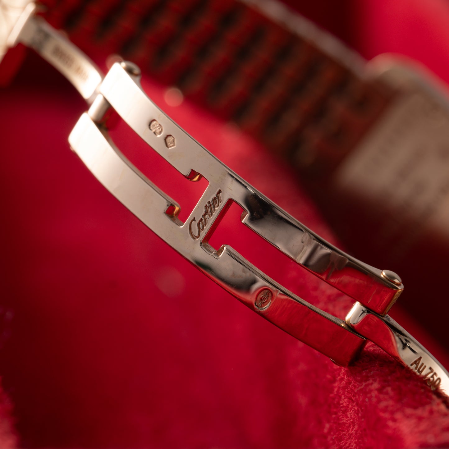 A close-up of the Cartier Tank Americaine's polished metal clasp features engraved Cartier logos and hallmarks, set against a blurred red backdrop.