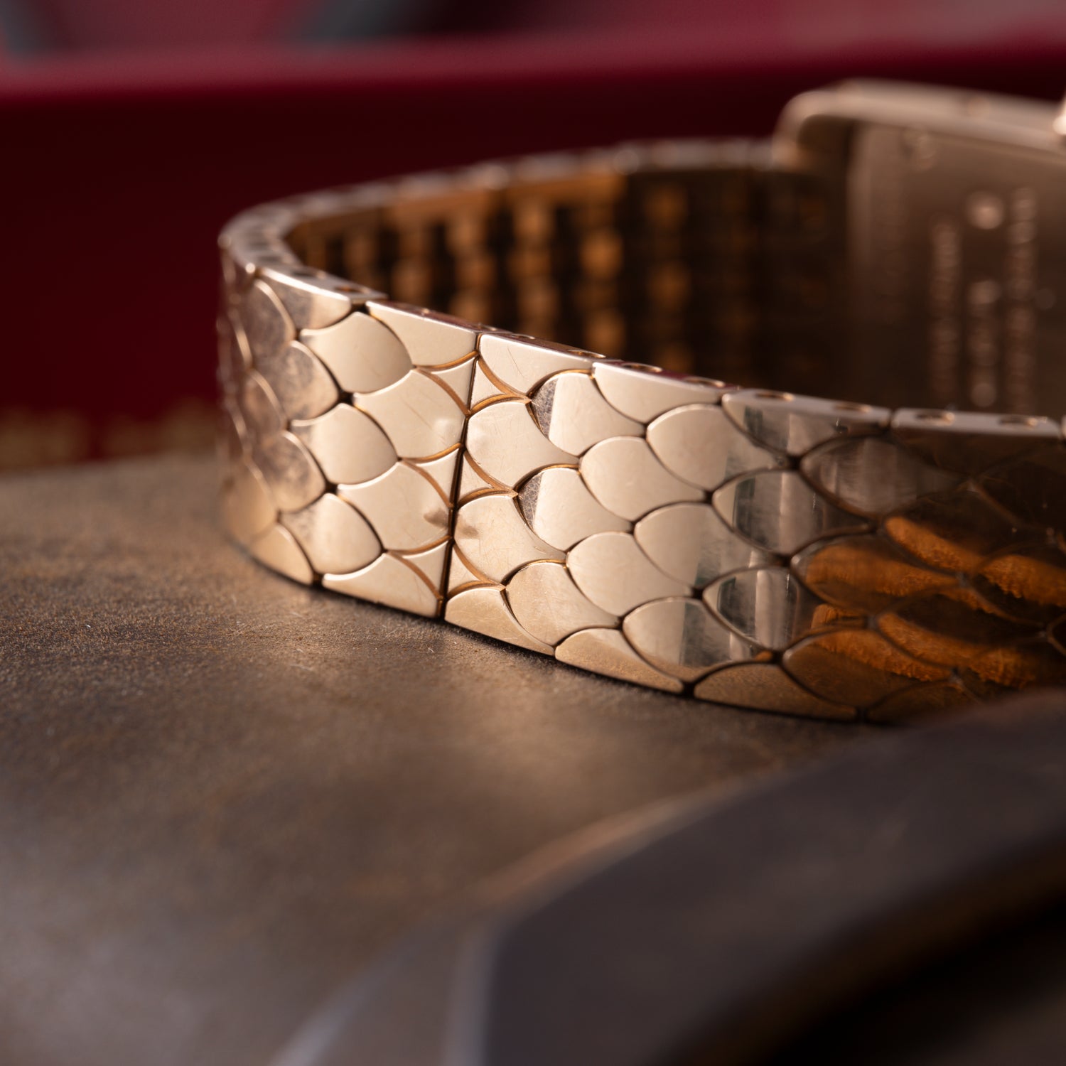 A close-up of a gold bracelet with a scale-like texture reflects light beside the Cartier Tank Americaine ladies 18K rose gold watch with a silver guilloché dial, set on a soft surface against a blurred red background.